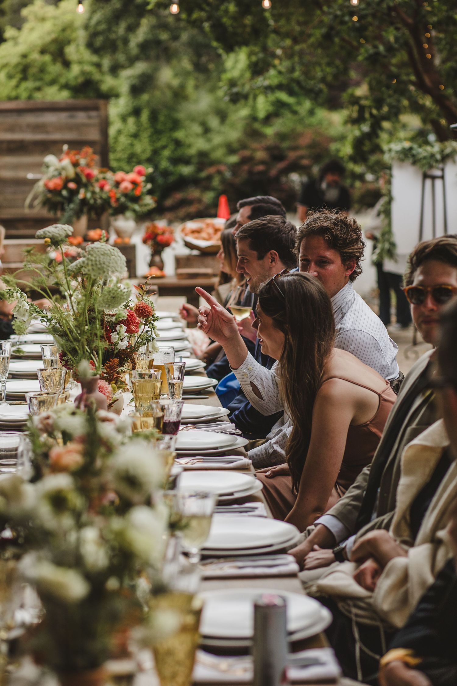 Guests seated at an elegant outdoor wedding reception table decorated with floral centerpieces and place settings.