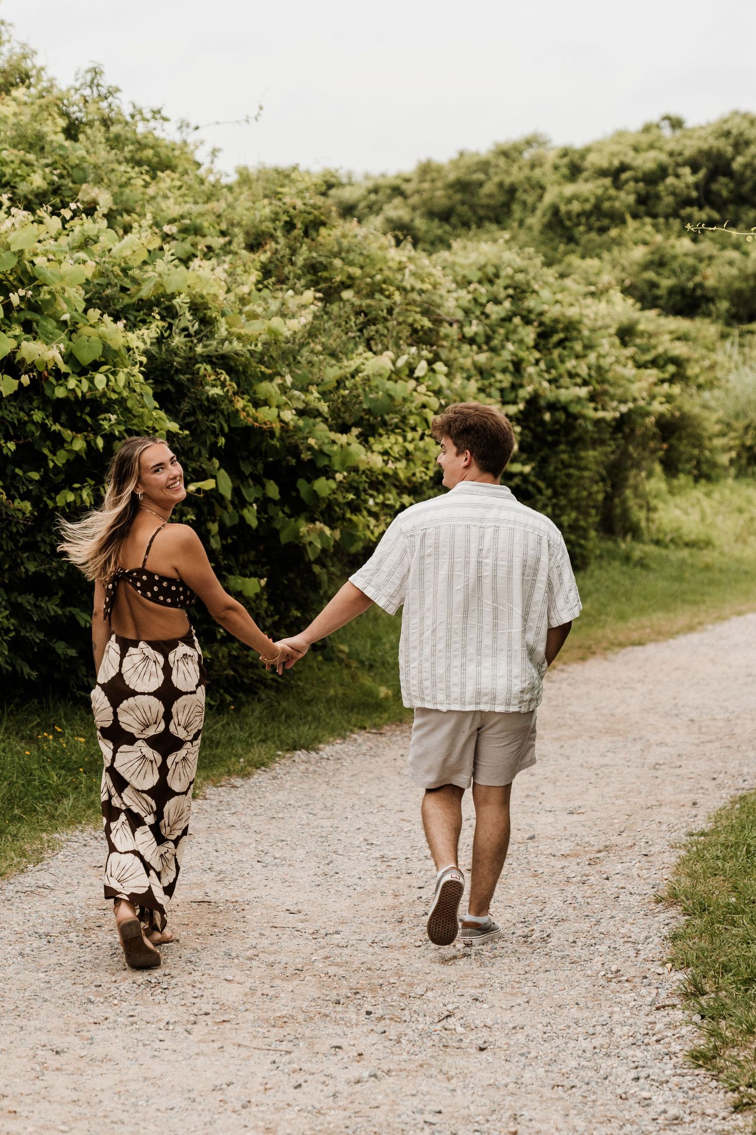 A couple walks hand in hand down a gravel path surrounded by lush green foliage on a sunny day.