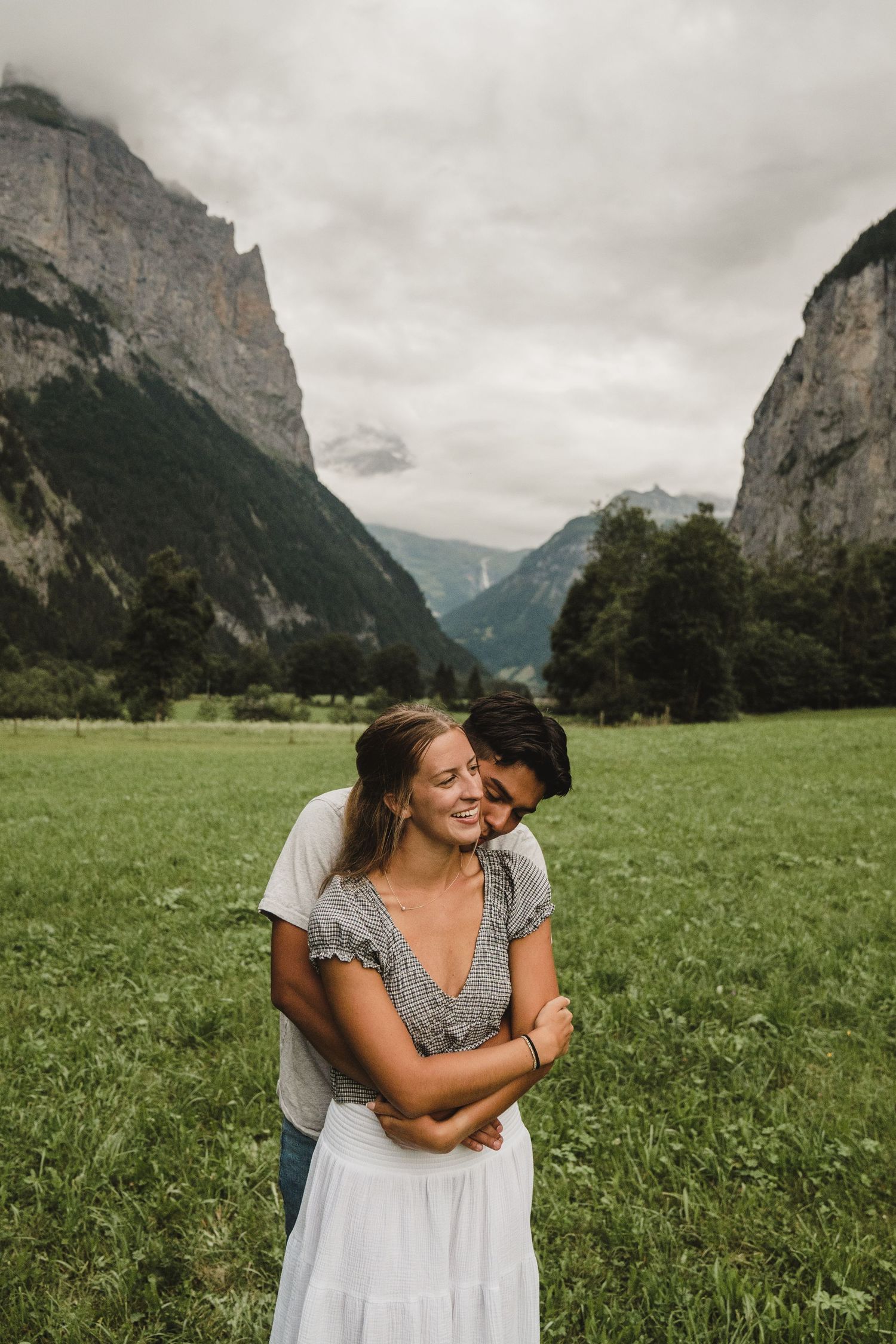 A couple embraces in a lush green meadow surrounded by towering mountain cliffs under an overcast sky in Switzerland.