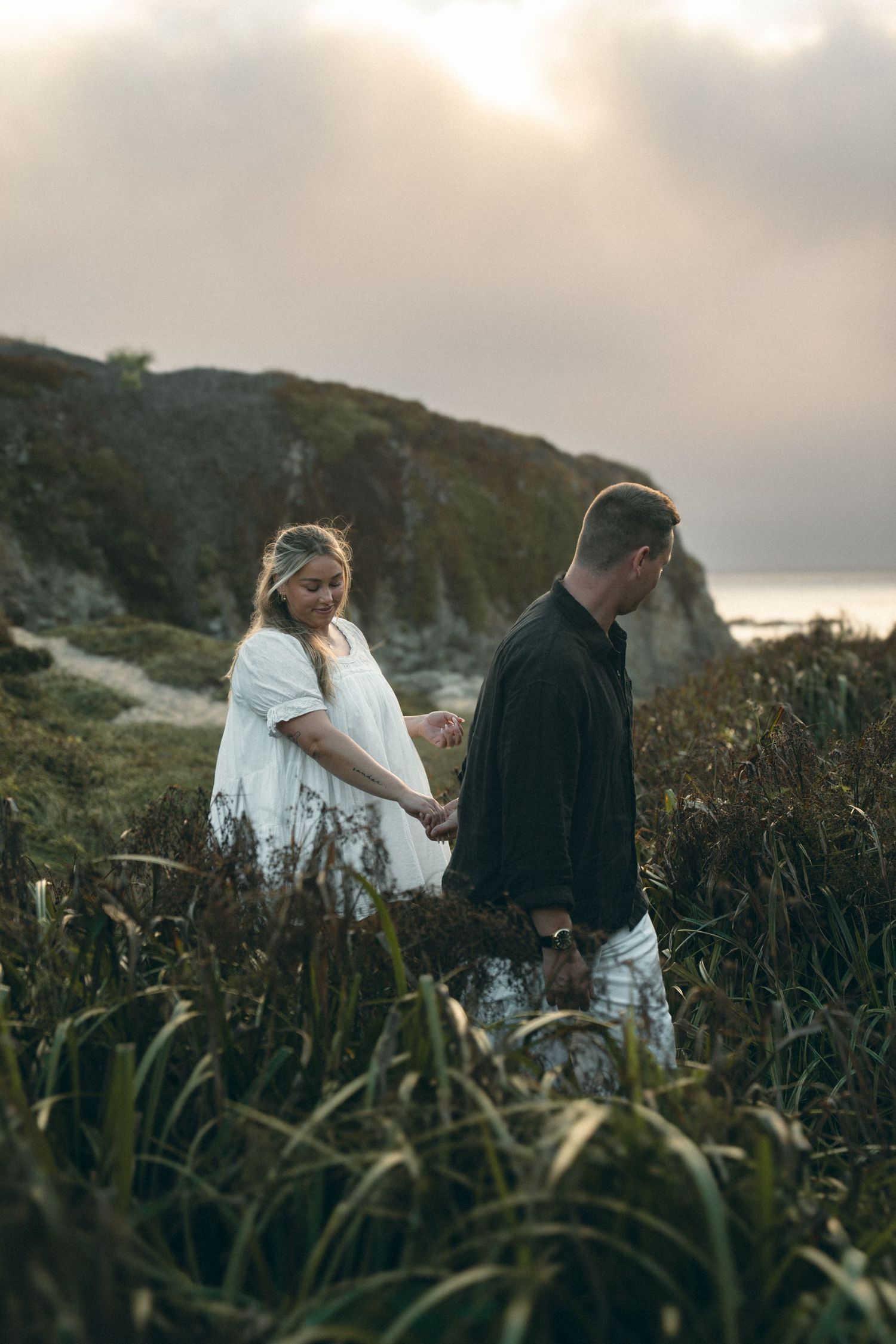 Two people stroll through tall grass along a rugged coastal hillside in moody evening light.