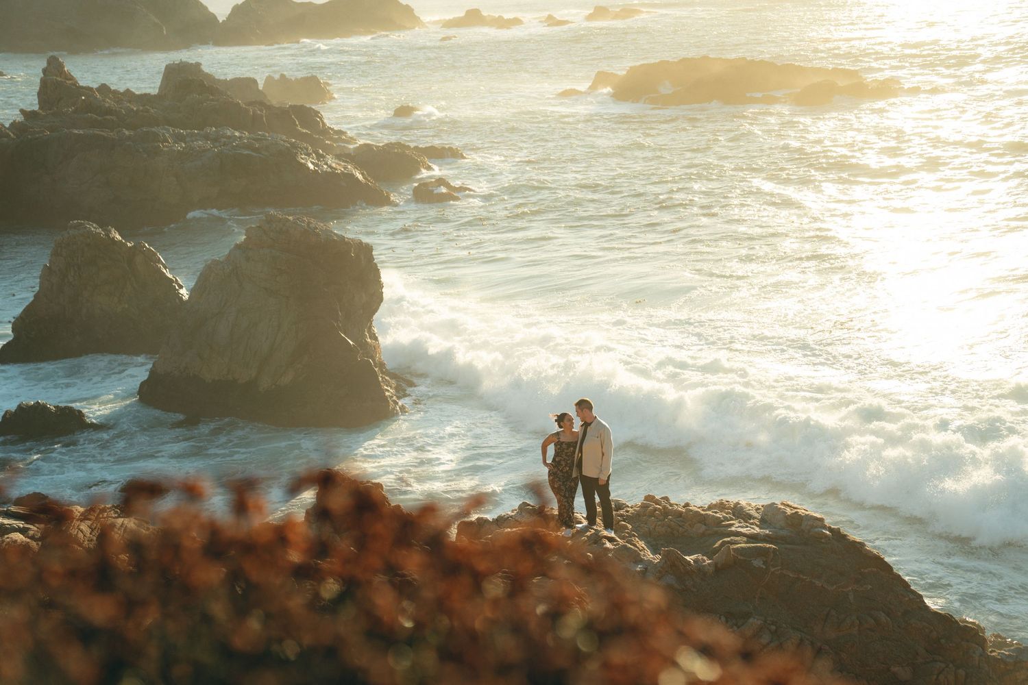 A group stands on rugged coastal cliffs at sunset with dramatic ocean waves crashing against rocks in the background.