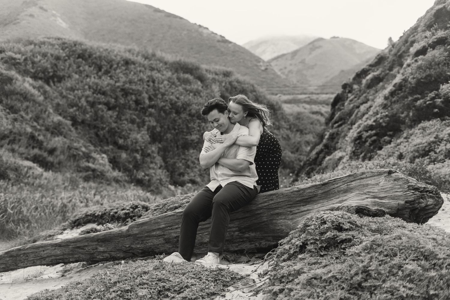 Couple embracing on driftwood log with scenic mountain hills in black and white outdoor photography.