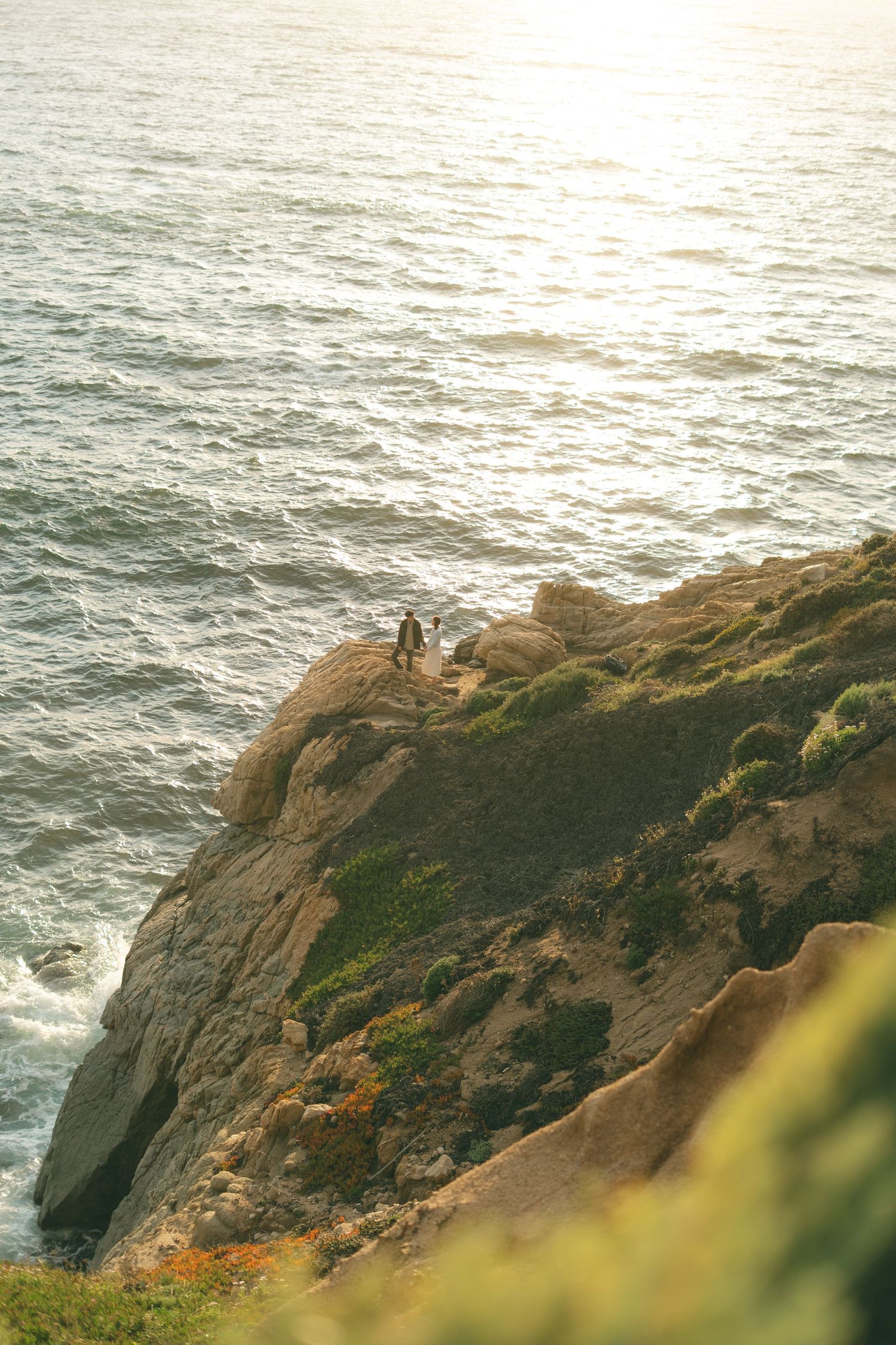 Two people standing on a rocky coastal cliff overlooking the shimmering ocean at golden hour.
