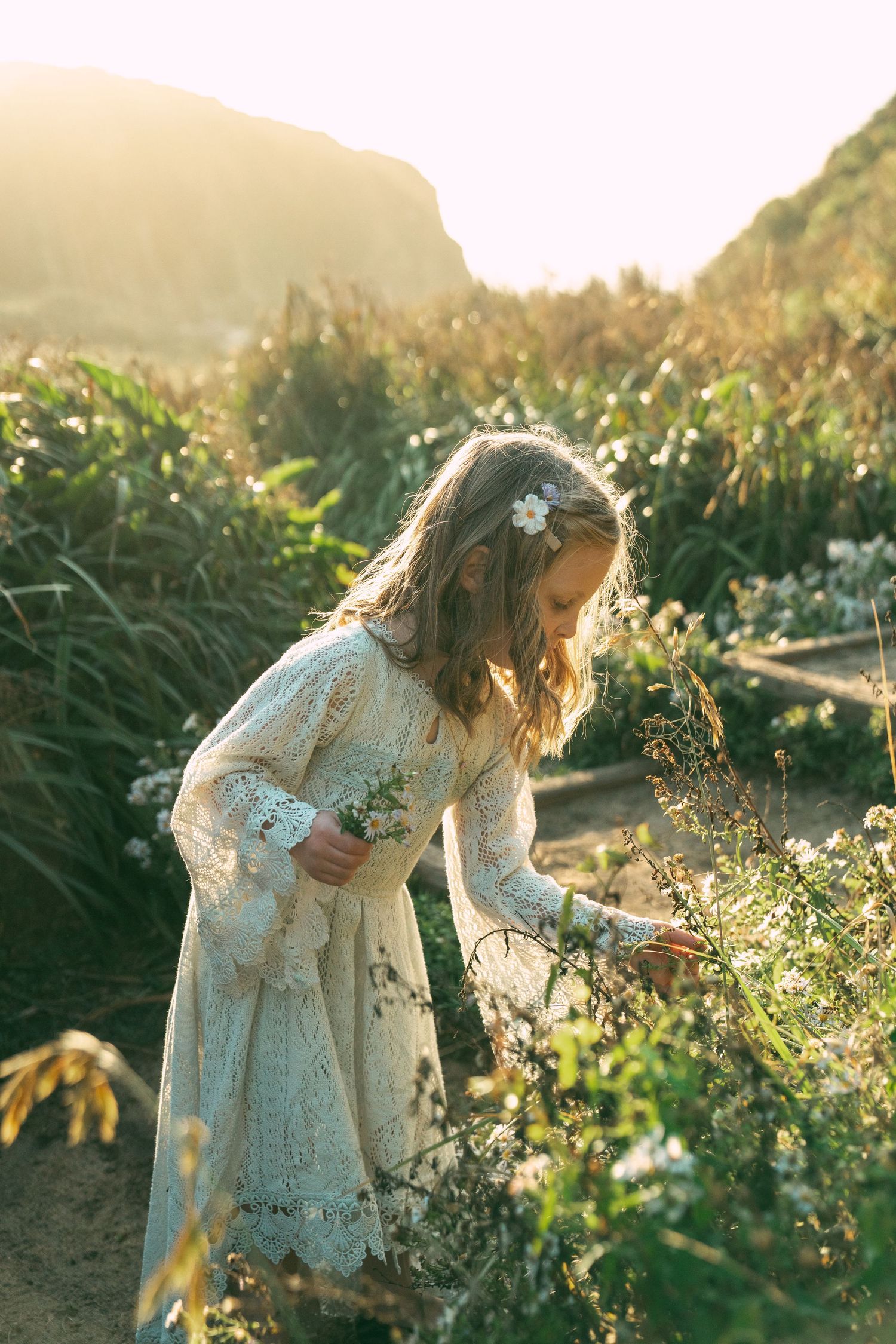 A dreamy scene of someone in a white lace dress picking wildflowers in a sunlit garden during golden hour.