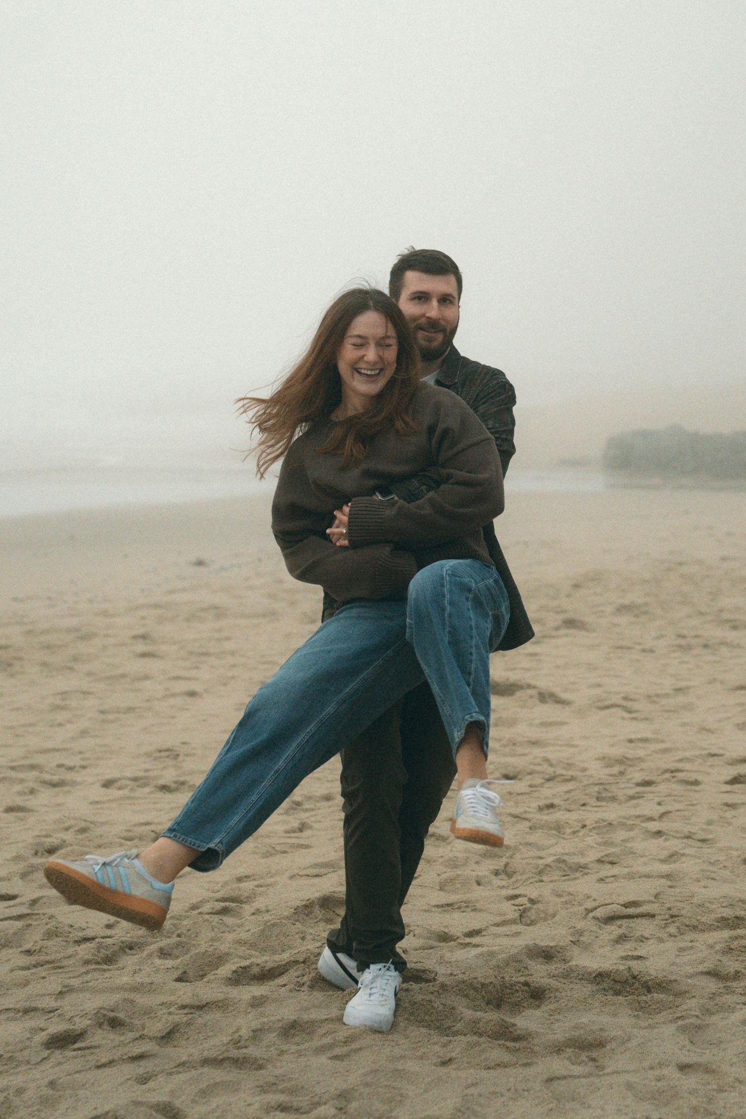 A couple playfully embracing on a foggy beach wearing casual attire.