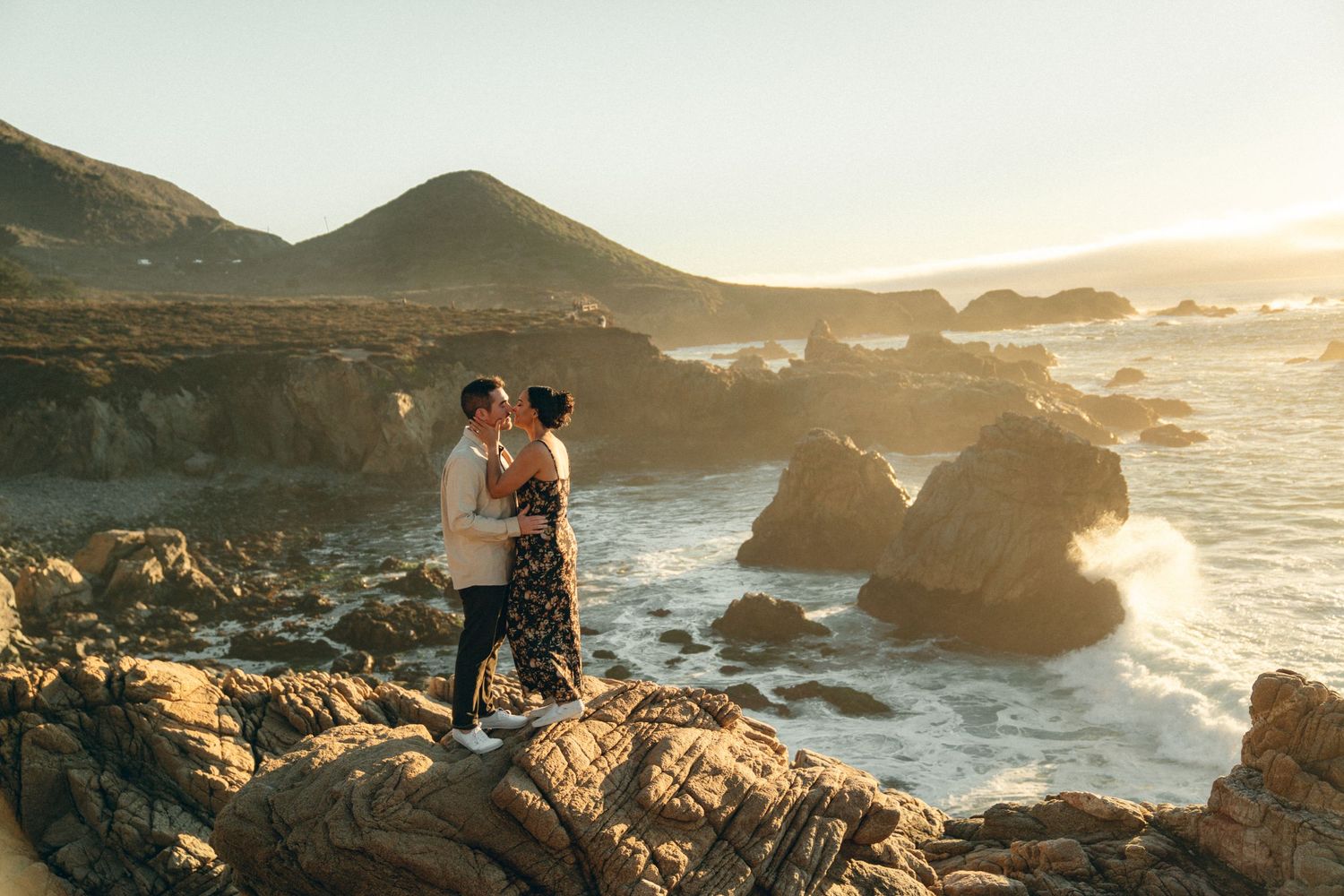 Couple embraces on rocky coastal cliffs at sunset with golden light illuminating the ocean waves below.