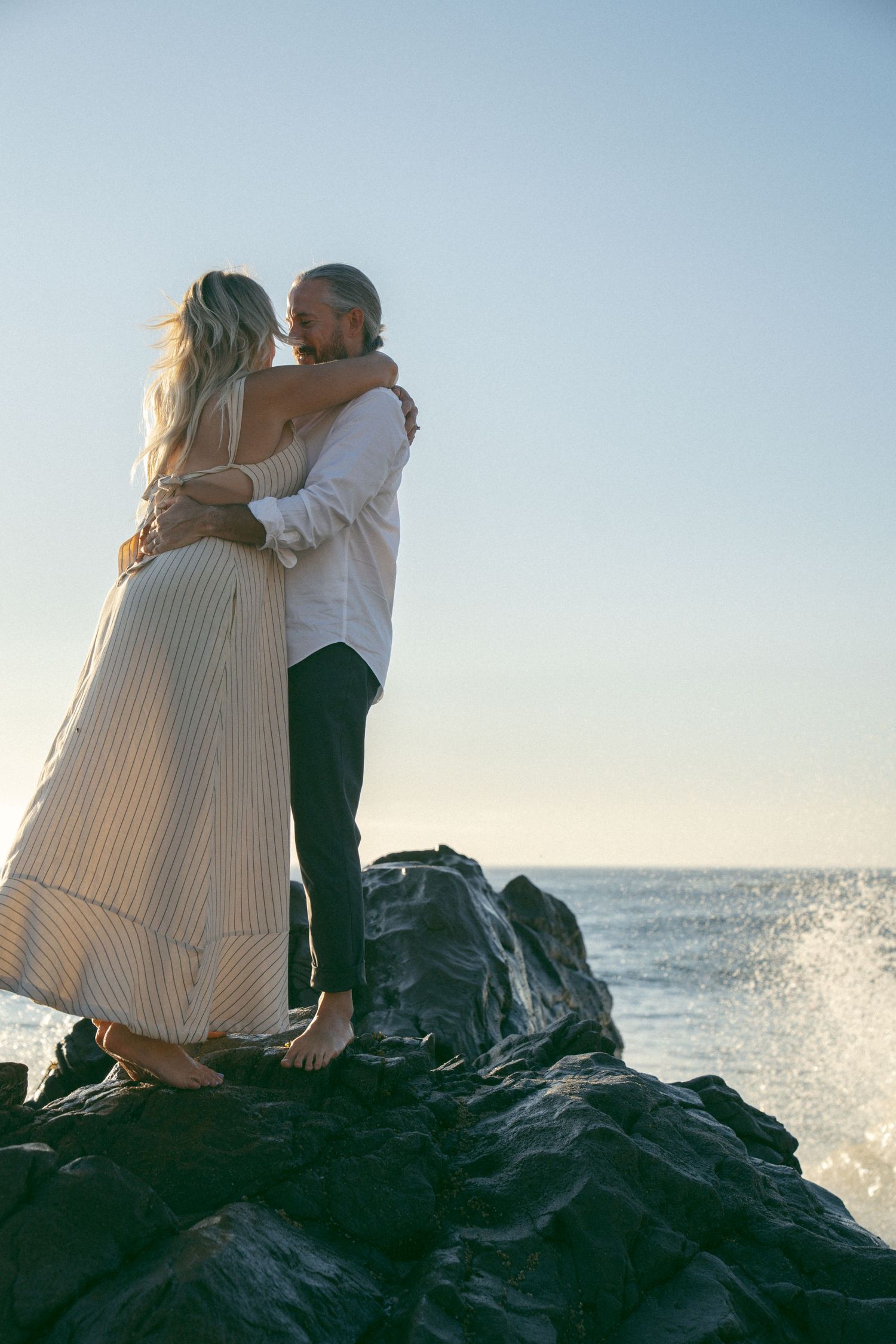 A couple embraces while sitting on coastal rocks as ocean waves splash behind them against a bright blue sky.