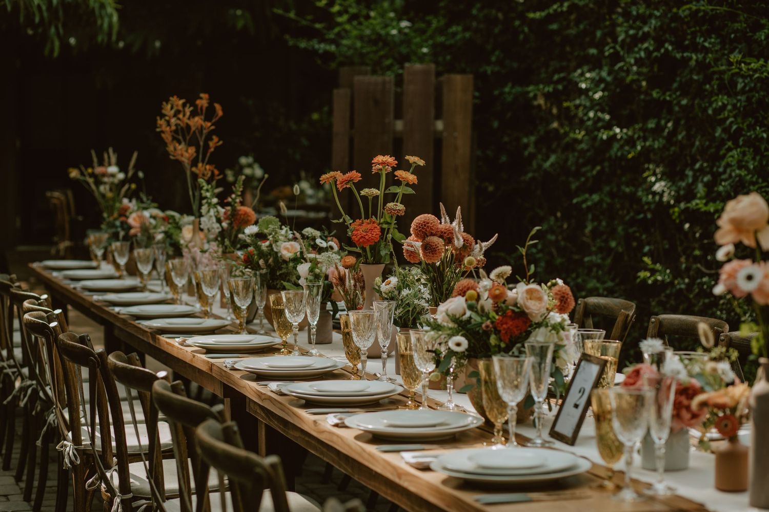A rustic outdoor table setting with floral centerpieces, wine glasses and place settings under a green forest canopy.