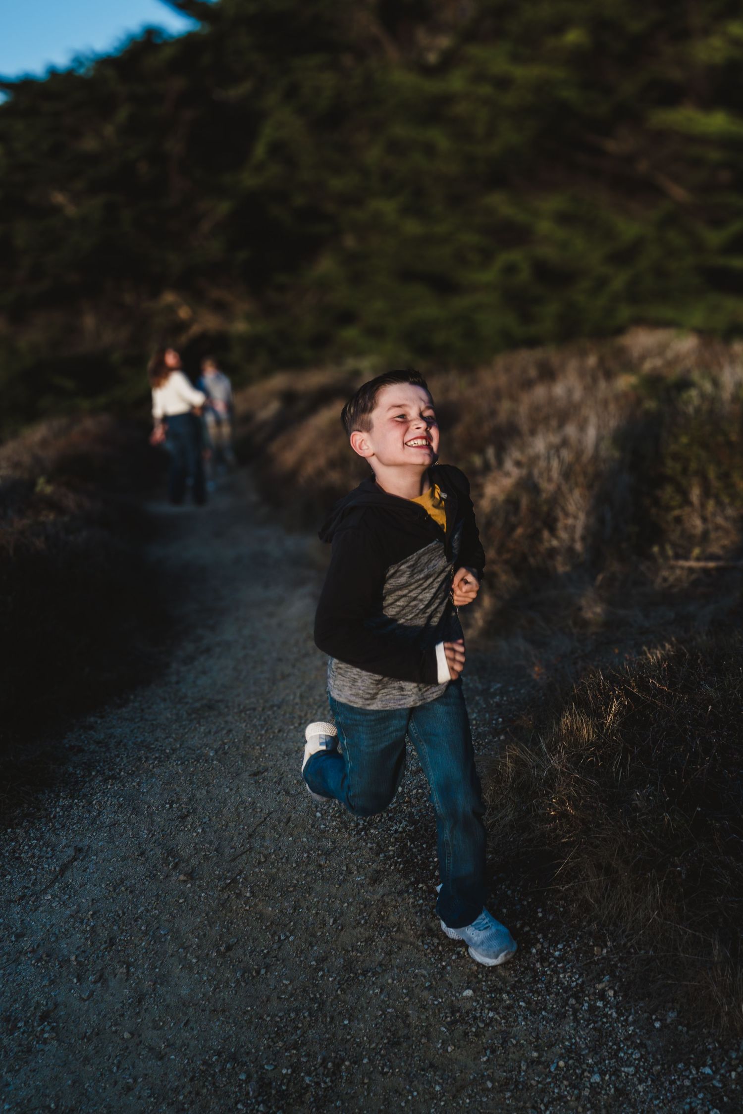 A child runs along a hiking trail at dusk while two figures can be seen in the background.