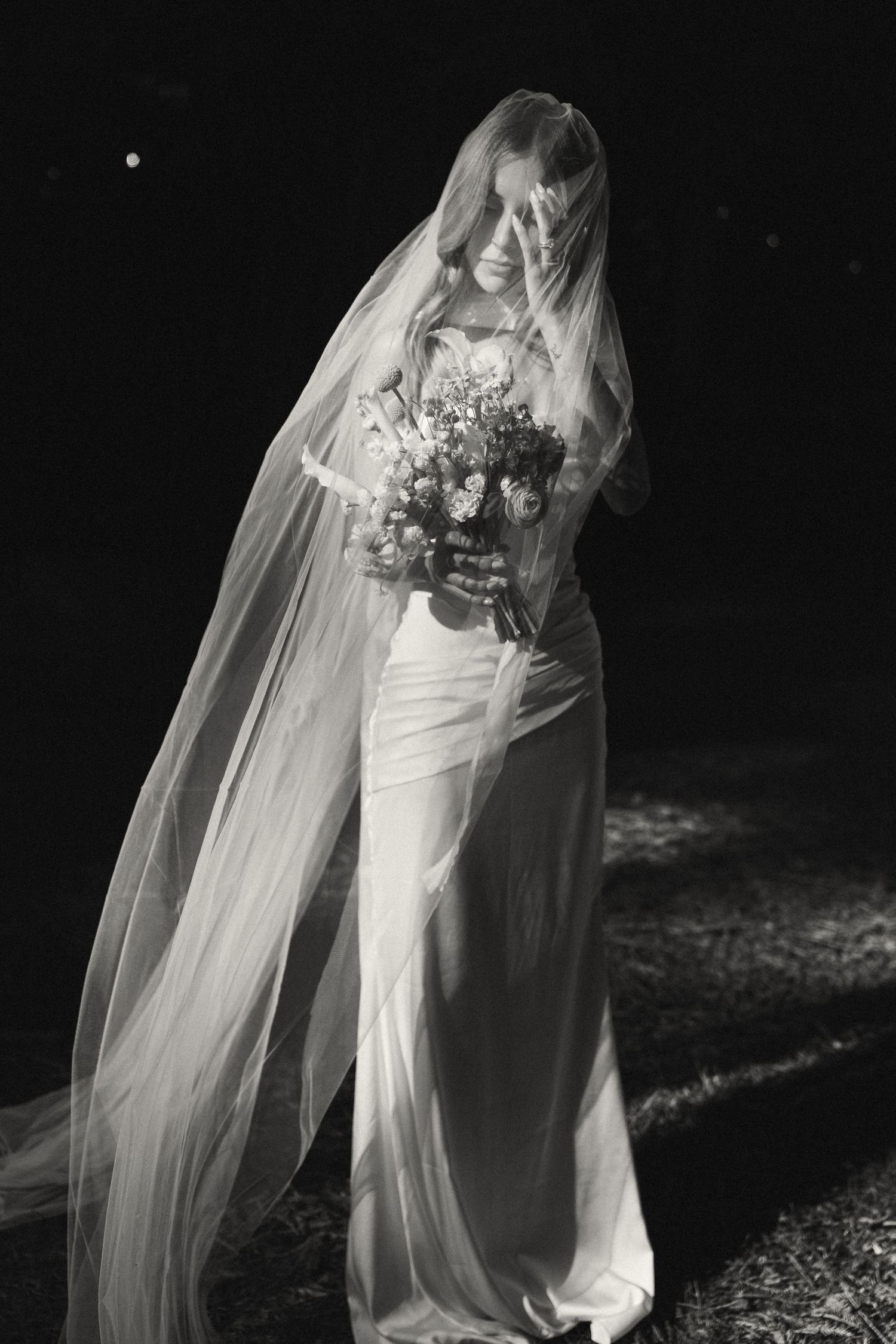 A bride in a flowing white dress and long veil holds a bouquet in this dramatic black and white photograph.