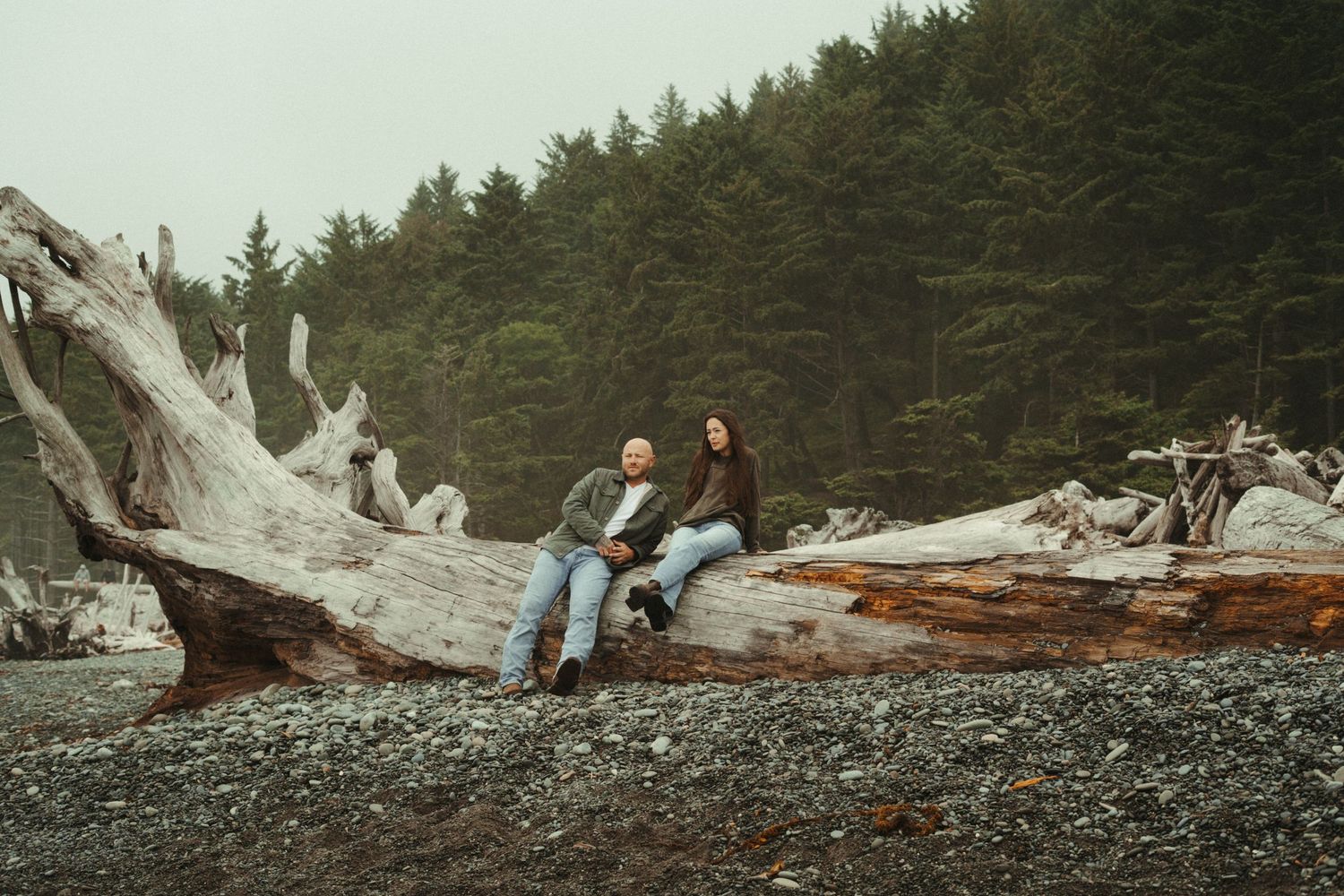 Couple sitting on driftwood logs on a rocky beach surrounded by evergreen forest on a cloudy day.