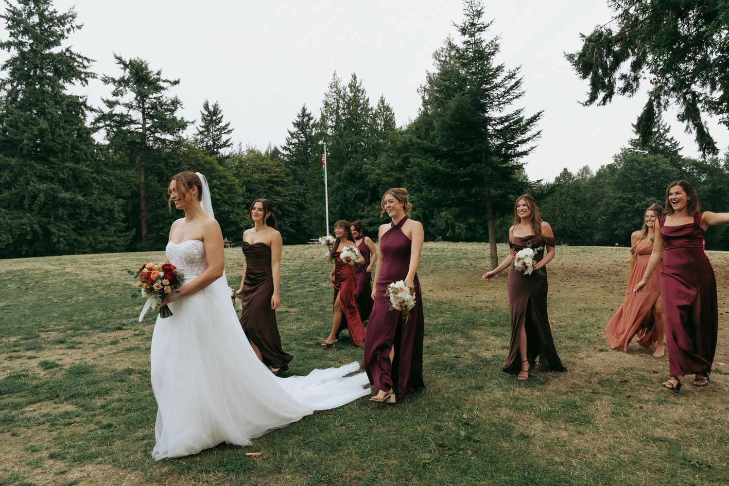 A wedding party stands on a grassy hillside in burgundy dresses and formal attire during an outdoor celebration.