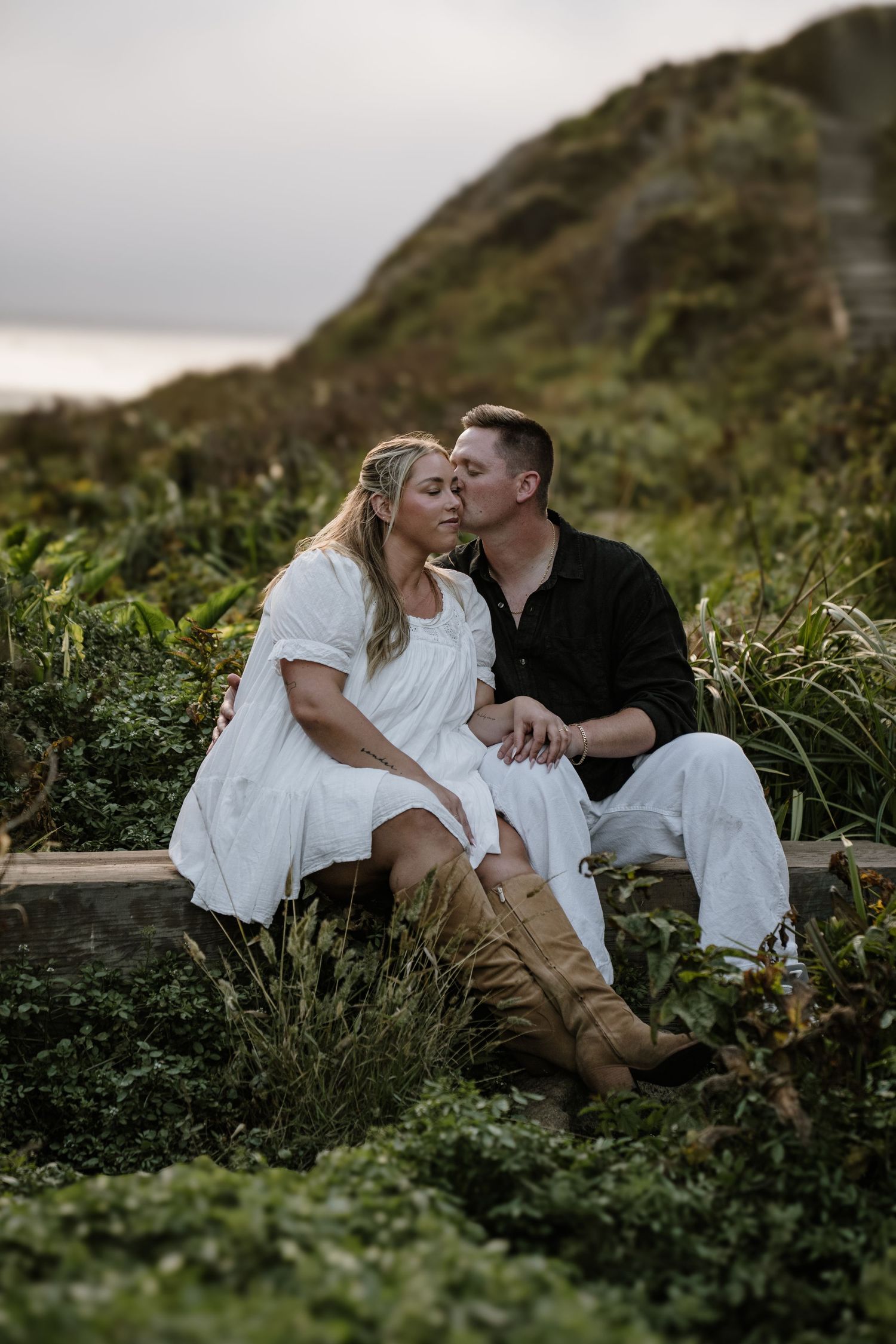 Romantic couple sitting together in tall grass on a hillside during a moody coastal photoshoot.