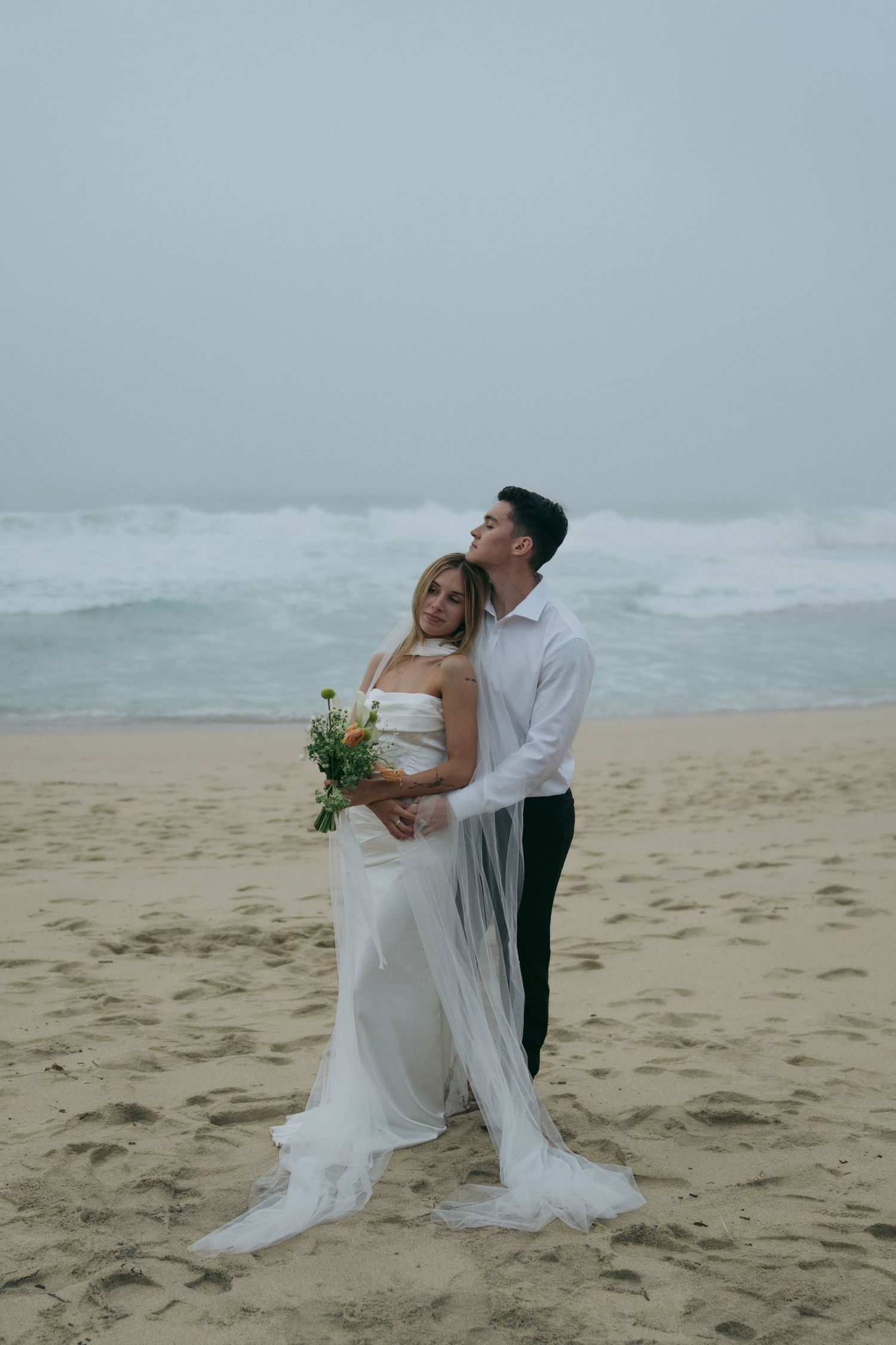 Romantic beach wedding photos with couple posing on sandy shore as waves crash behind them.