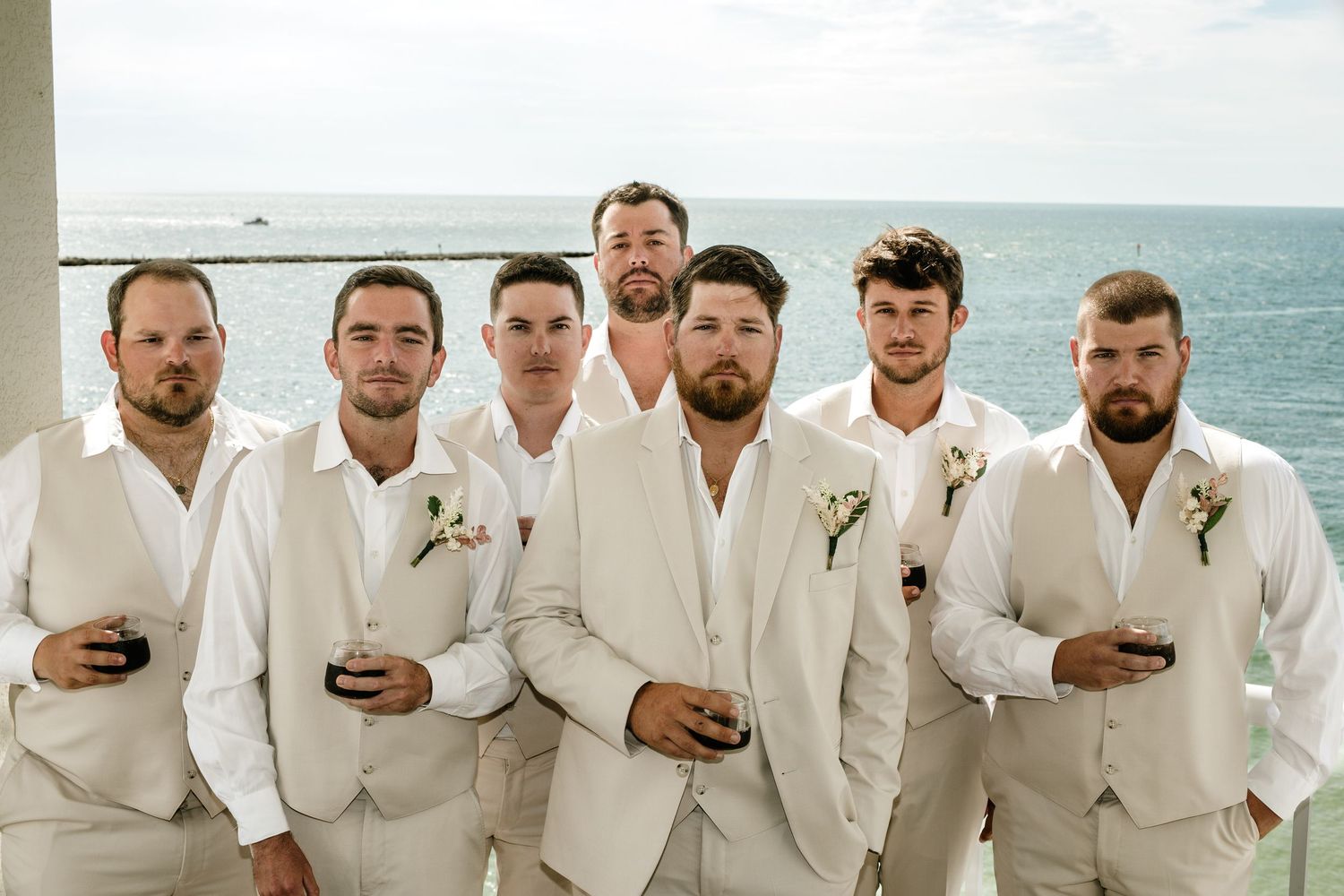 A groomsmen party poses together in crisp white suits with boutonnieres on a sunny beach overlook.