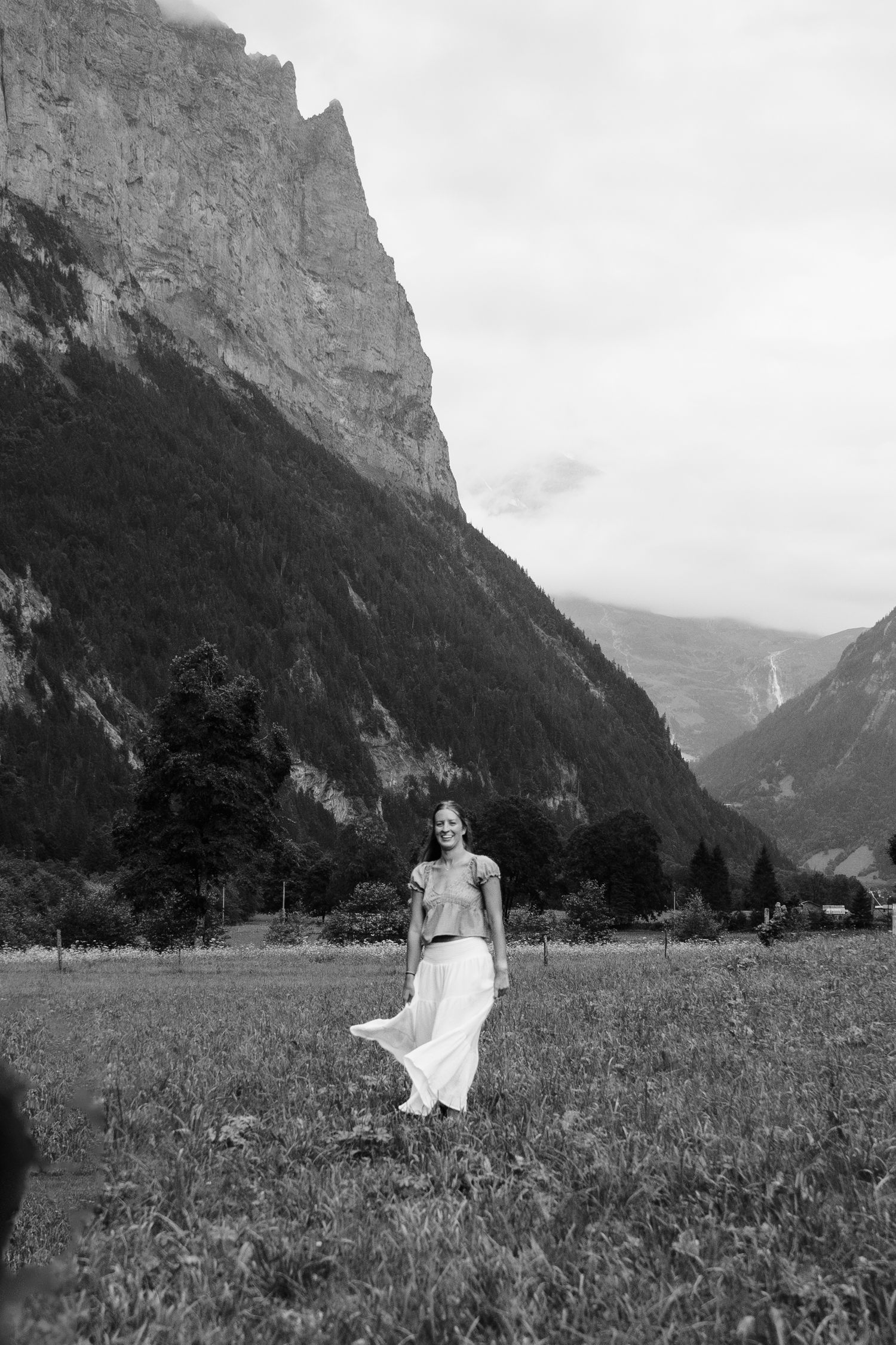 A figure in a white dress stands in a meadow with dramatic mountain peaks rising in the background in black and white.