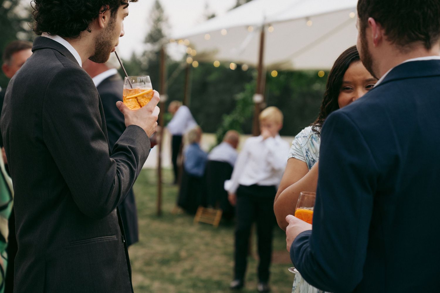 People enjoying drinks at an outdoor social gathering with string lights and greenery in the background.