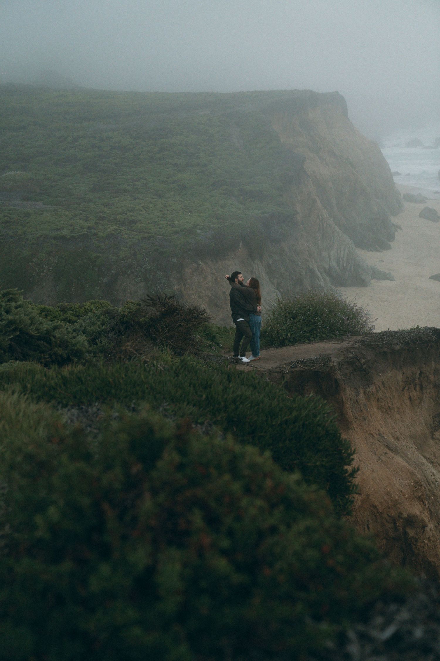 A misty coastal scene with steep cliffs overlooking the ocean where two people embrace on a foggy day.