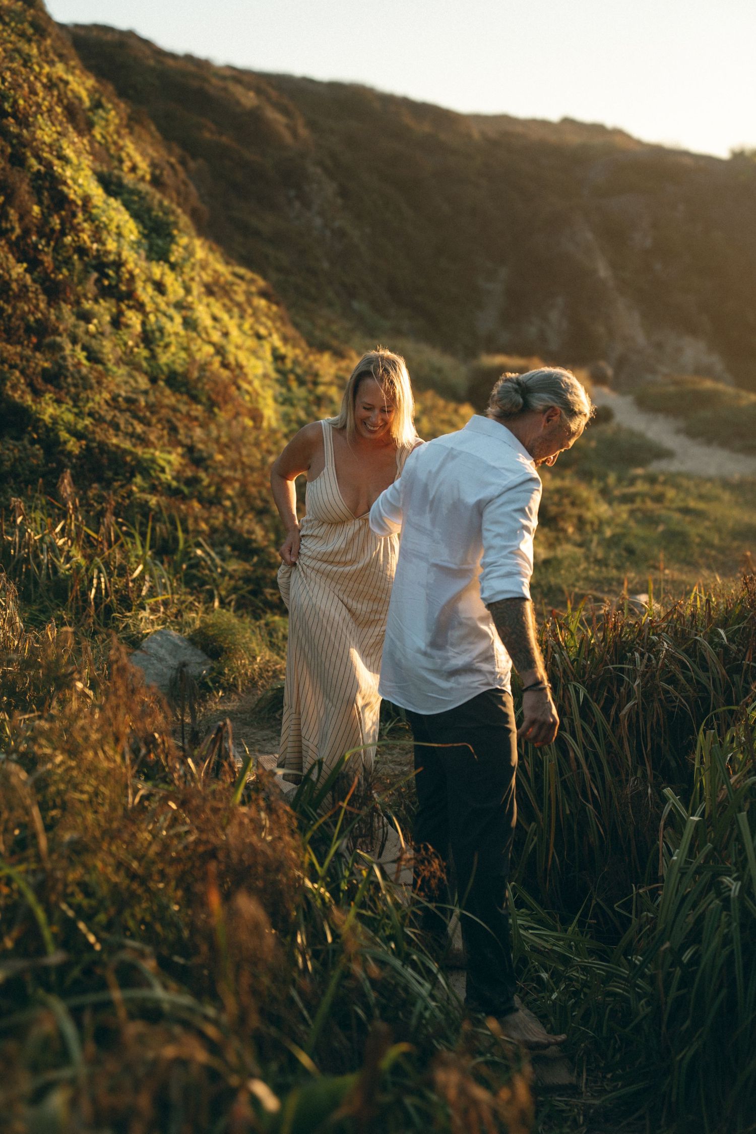 A couple walks together through tall grass on a hillside during golden hour sunset.