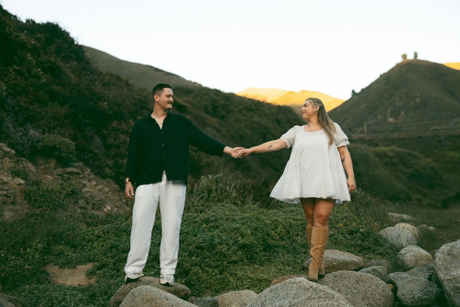 Two people walk together along a scenic mountain trail at sunset while wearing casual outdoor attire.