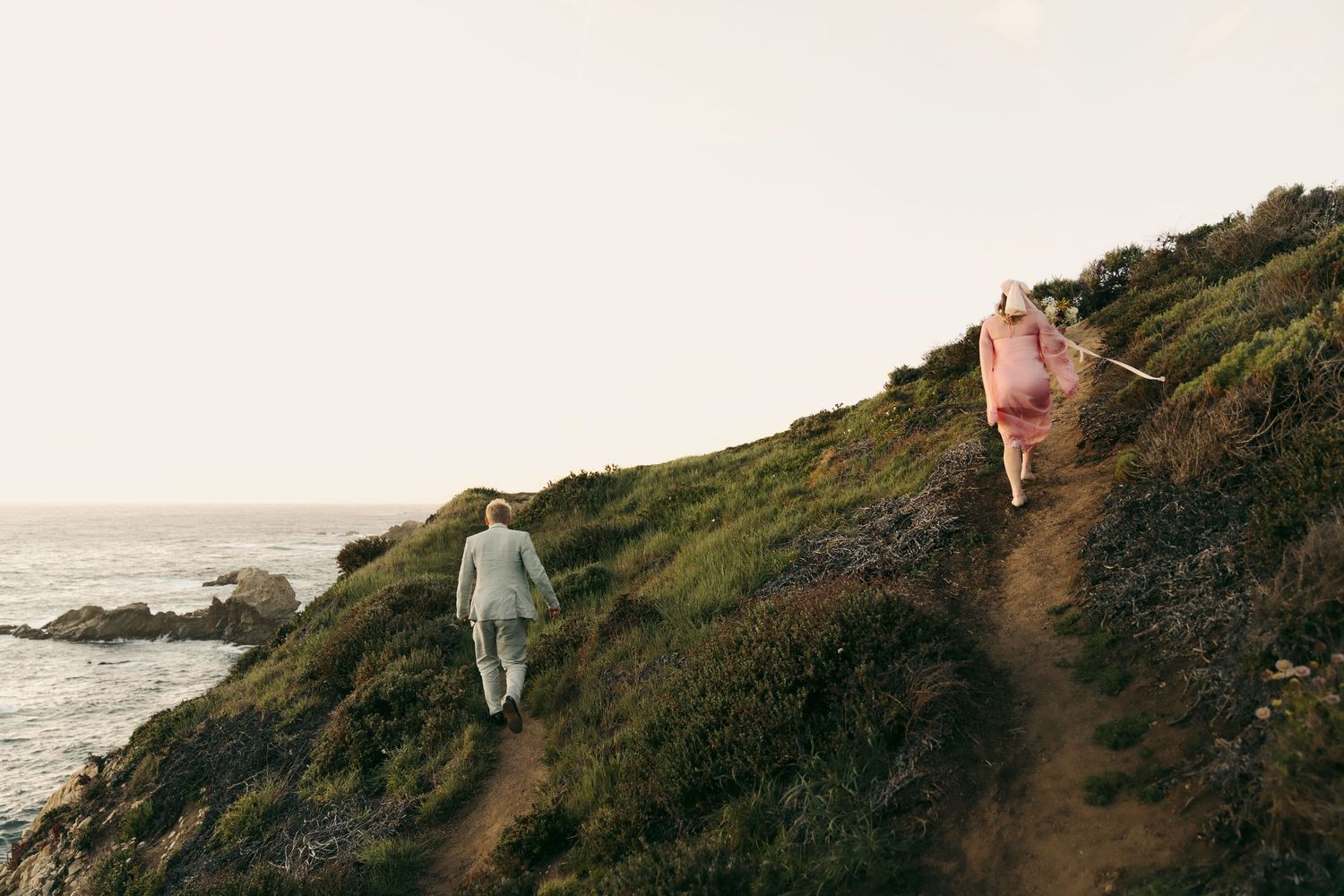 Couple walking up a coastal hillside path, man in gray suit and woman in pink dress, with ocean views.