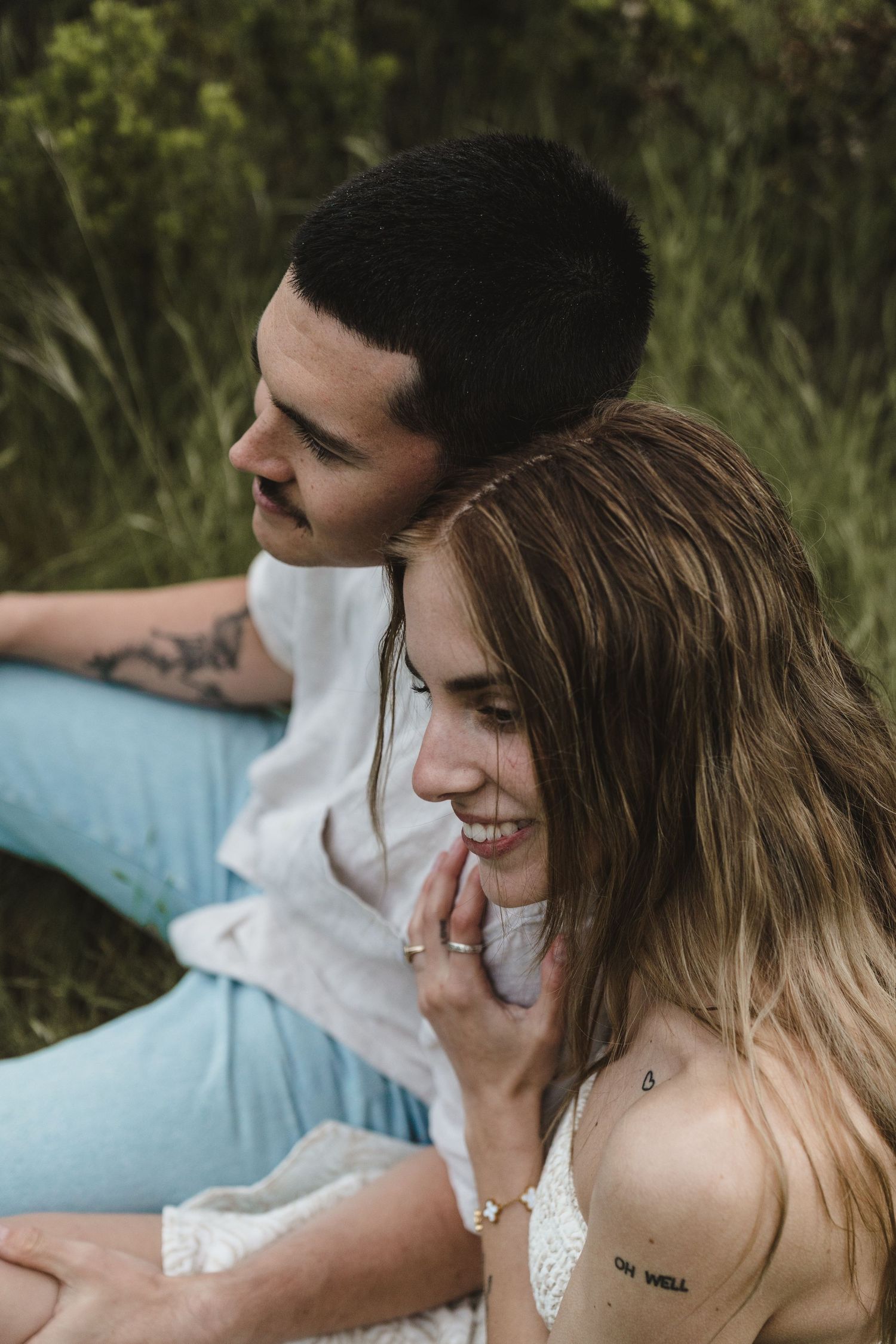 A couple sits together on grass wearing light colored clothing while sharing a candid laughing moment in Big Sur California.