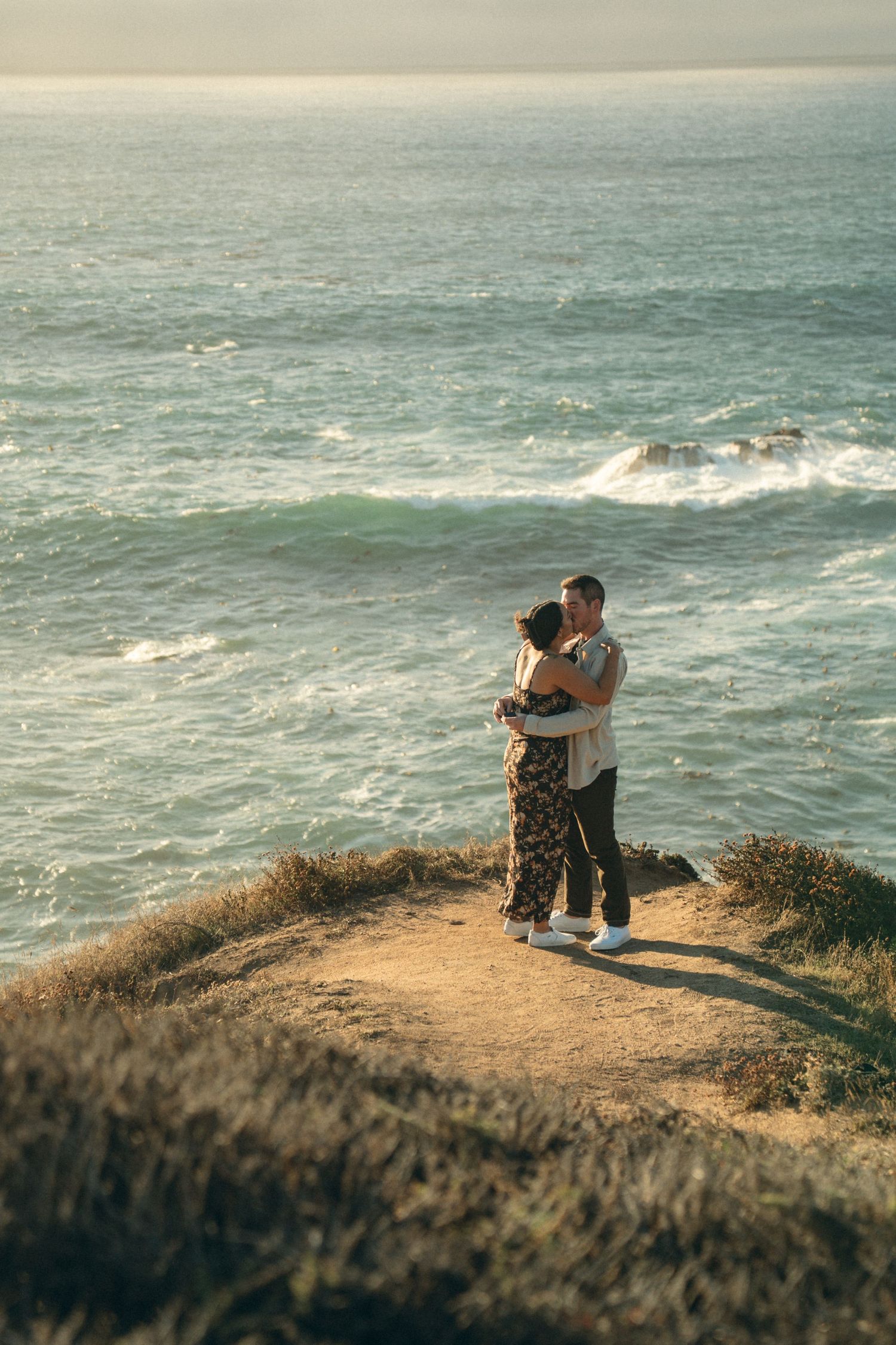 A couple embraces while standing on a coastal cliff overlooking the ocean during golden hour.