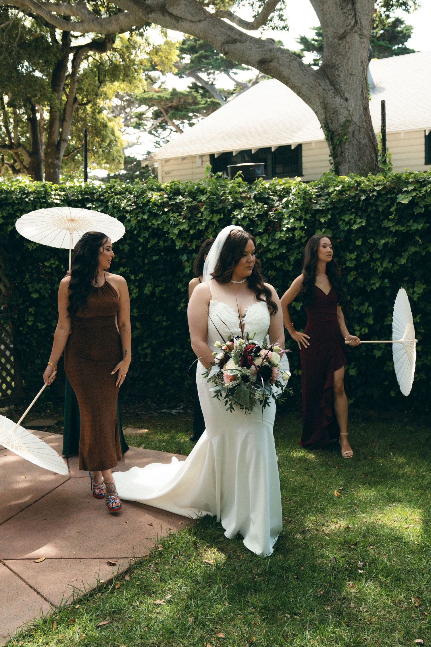 A Carmel-By-The-Sea wedding party poses playfully in a garden setting with white parasols and flowing dresses.