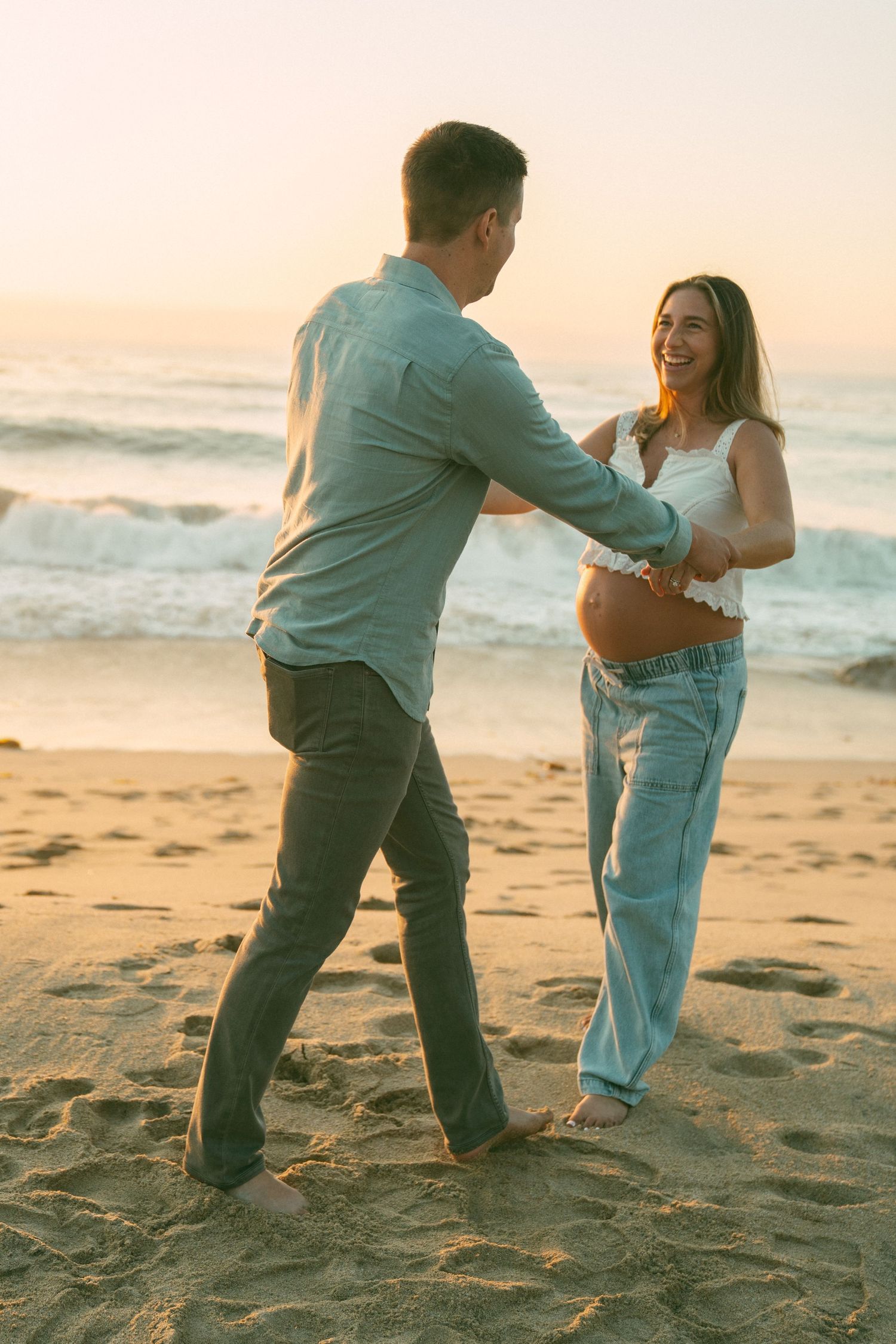 A couple playfully interacts on a sandy beach at sunset with waves in the background.