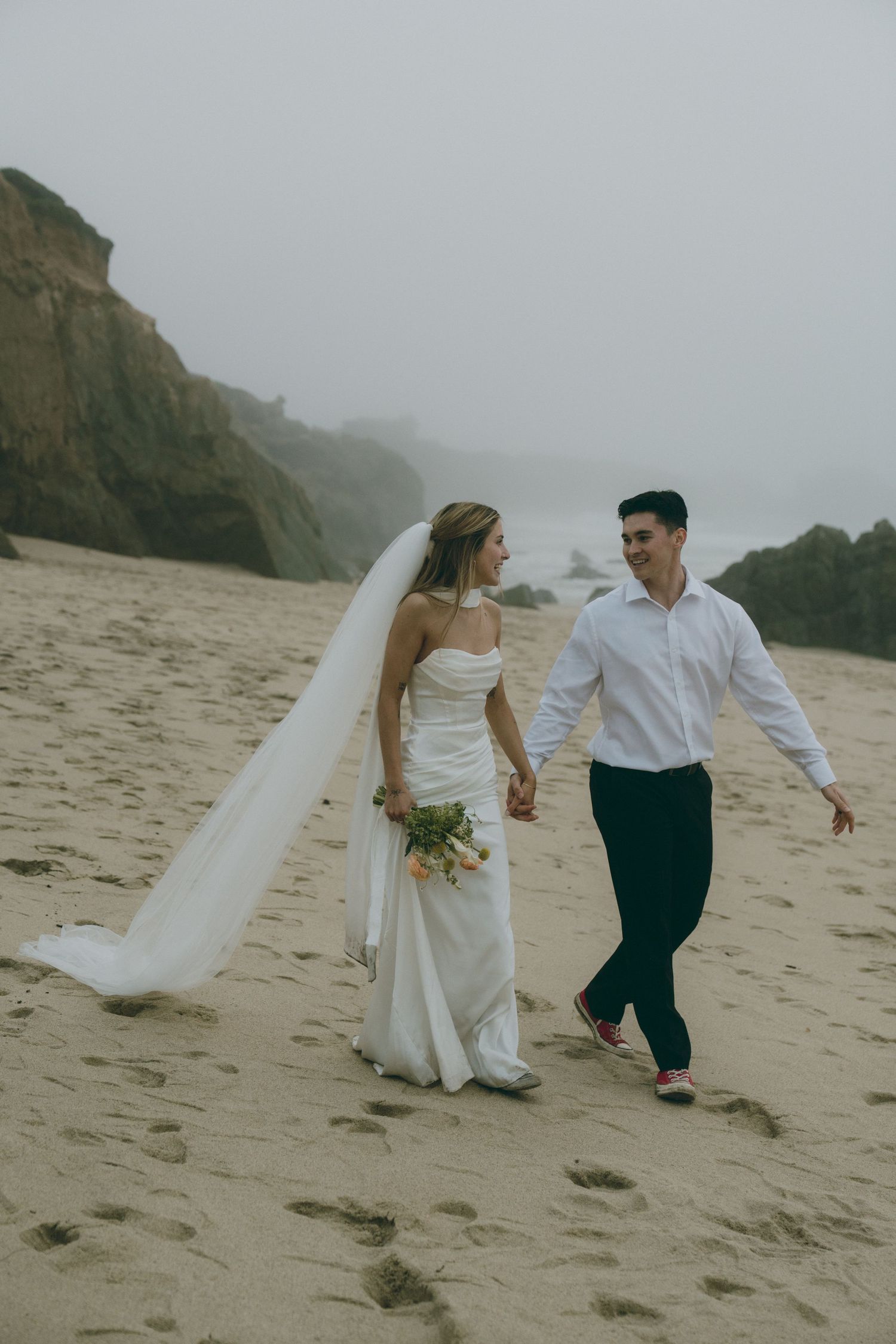 A couple walking and holding hands on a foggy beach with mountains in the background.