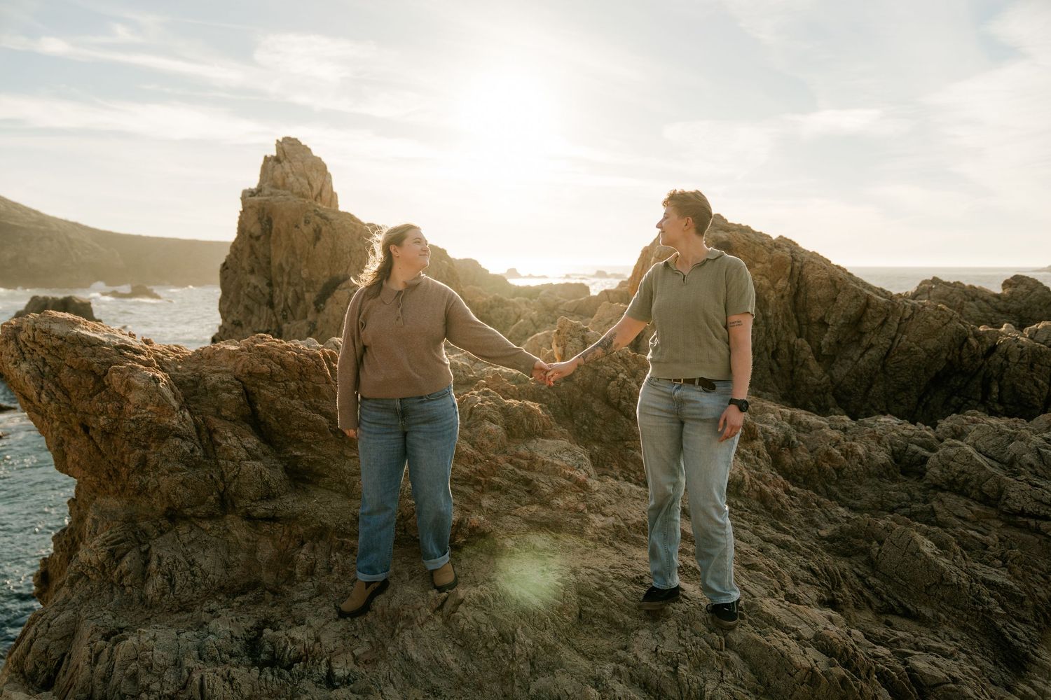Two people silhouetted against sunset sky on rocky coastal outcrop with ocean in background.
