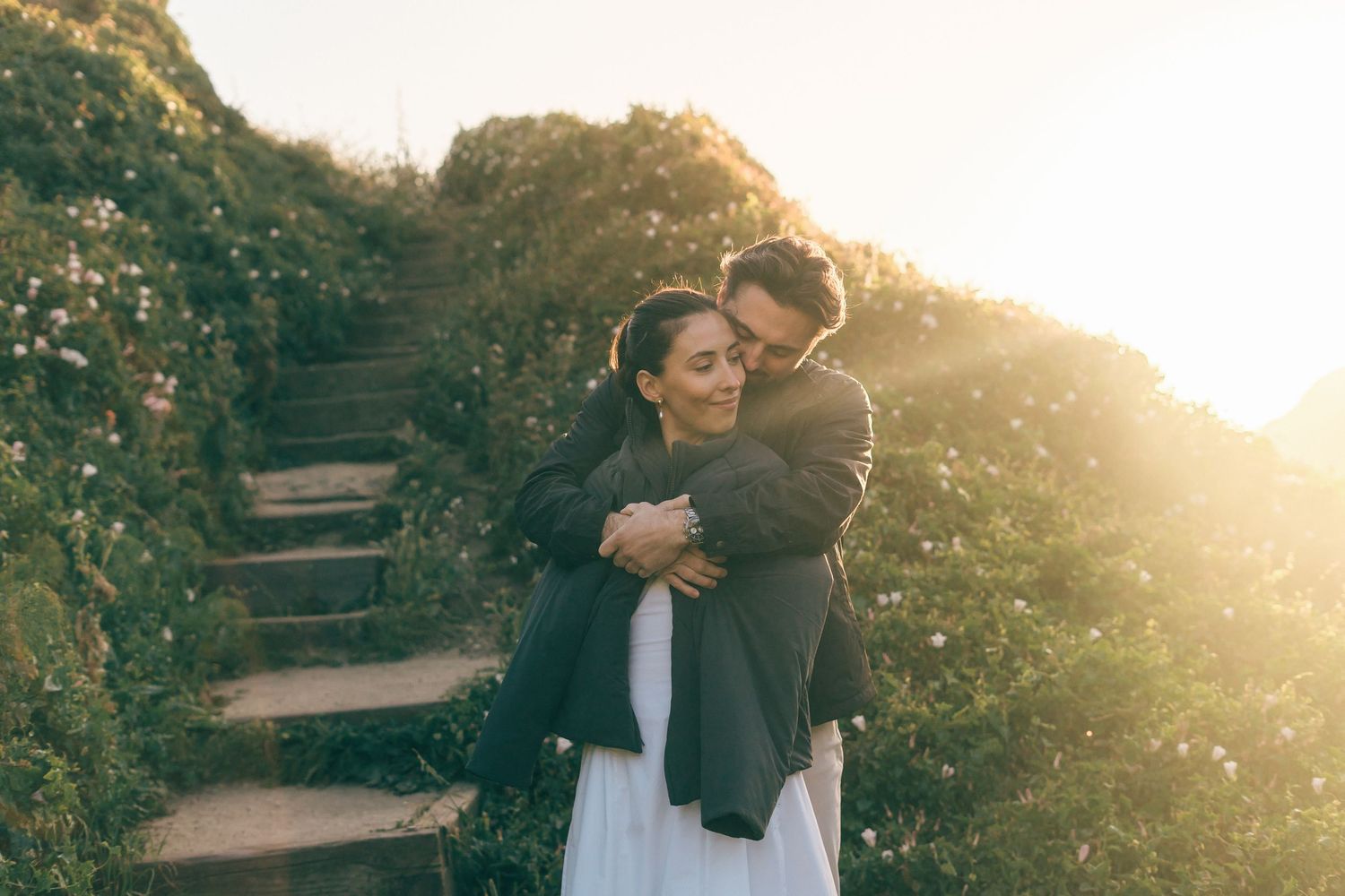 Couple embracing on stone steps surrounded by lush greenery in golden sunset light.