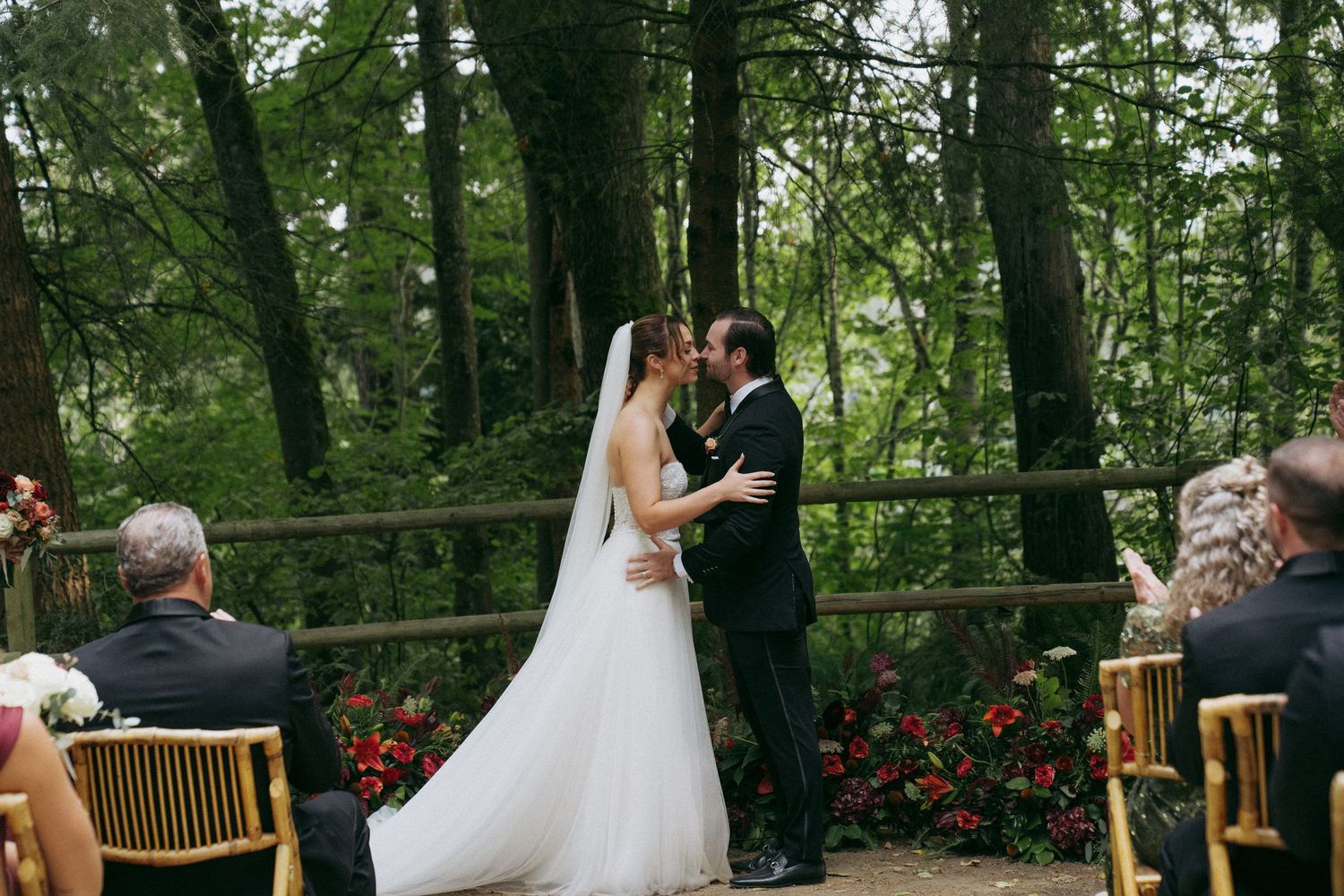 A couple shares a romantic kiss during their outdoor wedding ceremony surrounded by lush green forest and wooden benches.