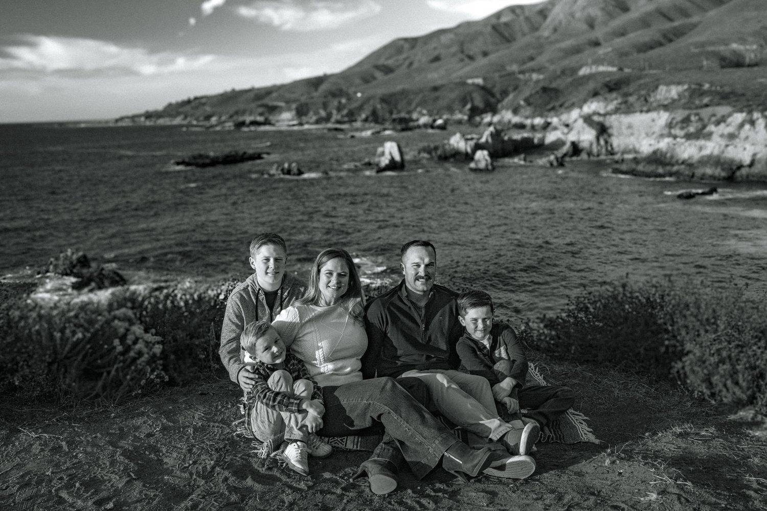 Black and white photograph of people gathered on a scenic coastal overlook with rugged cliffs and ocean views.