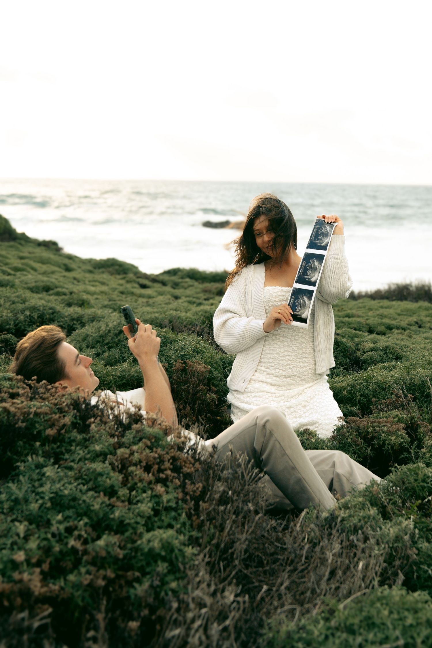 Couple at rocky seaside, woman in white dress holding ultrasound photos while man photographs her, pregnancy announcement.