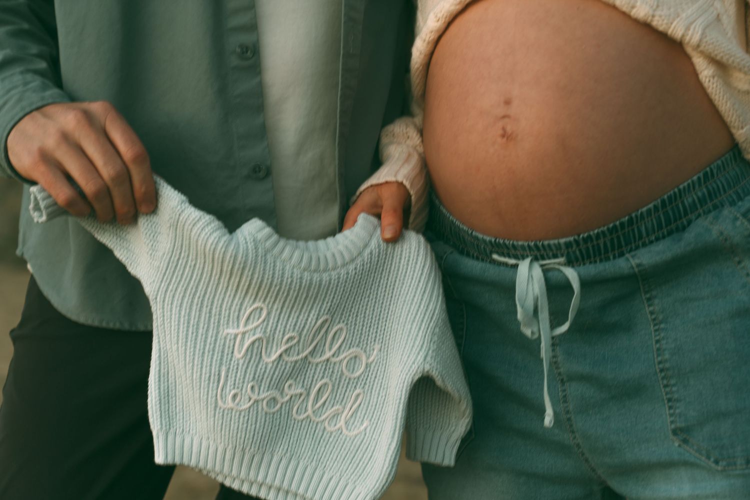 Pregnant woman holding a white 'Hello World' knit sweater next to her bare baby bump in soft neutral tones.