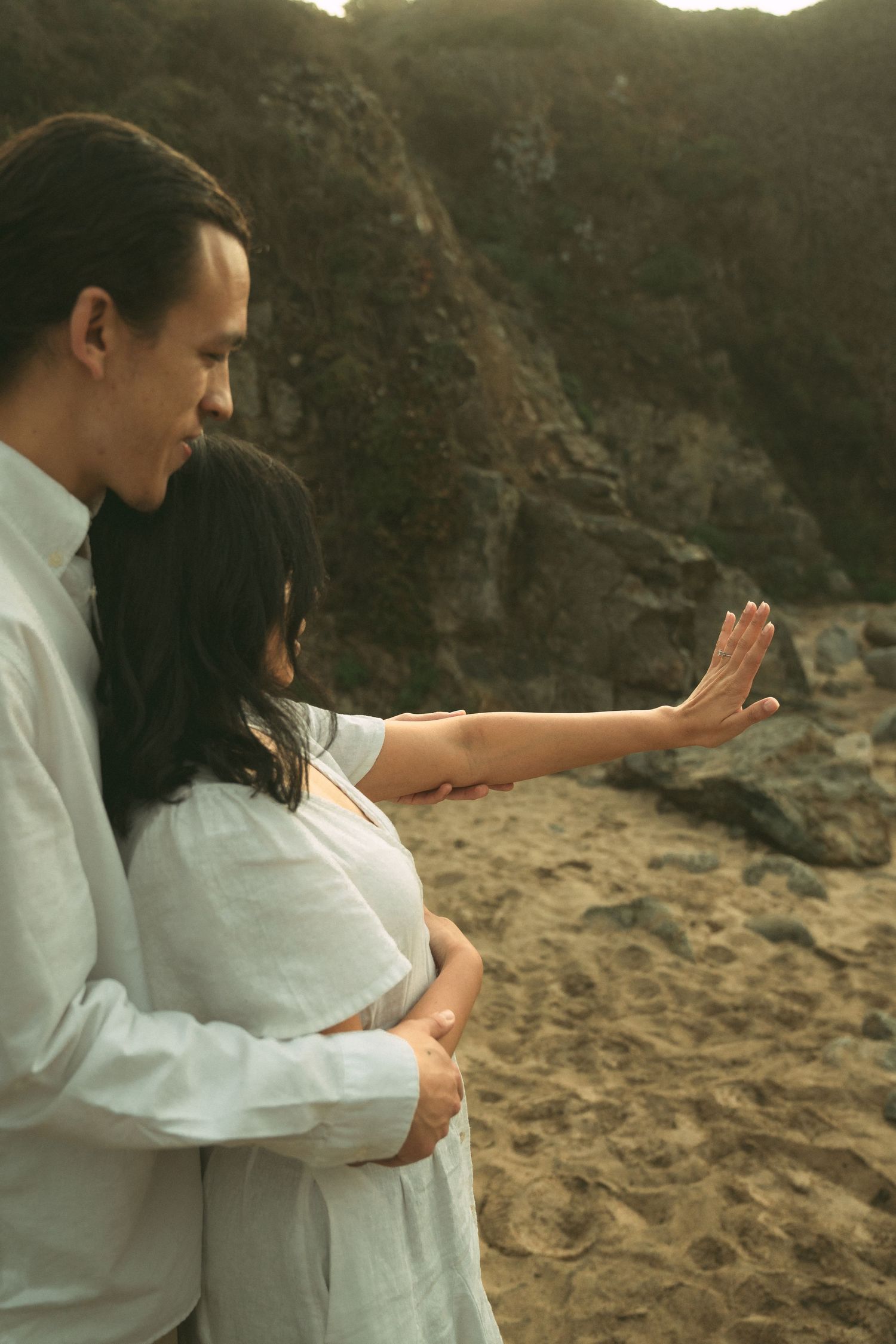 A couple in white clothing embraces tenderly on a rocky beach at sunset.