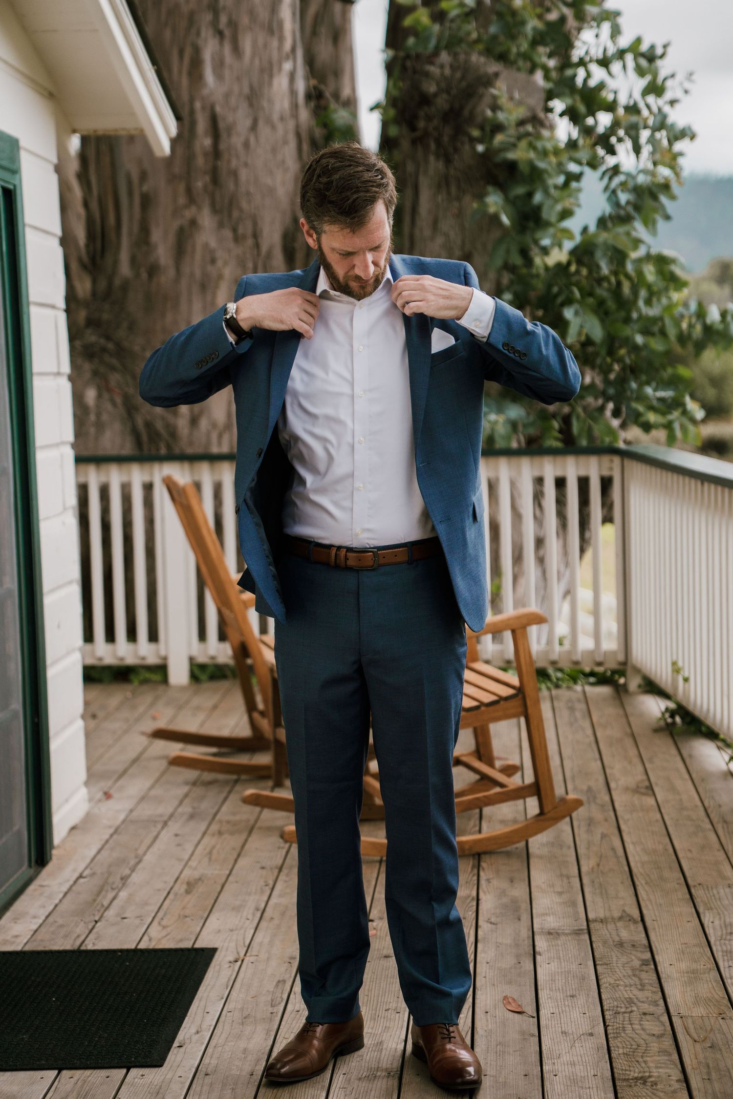 Close-up details of groom's formal navy suit and brown leather shoes on rustic wooden porch.