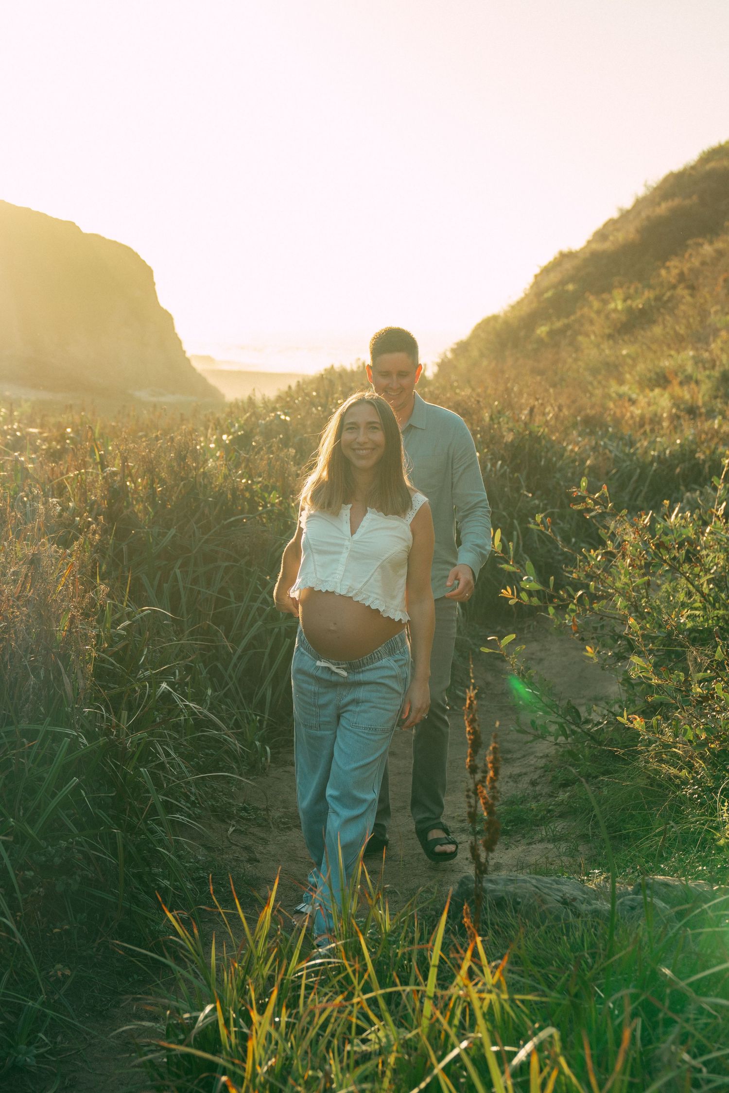 Two people walk through a lush green field with mountains and ocean views at sunset.