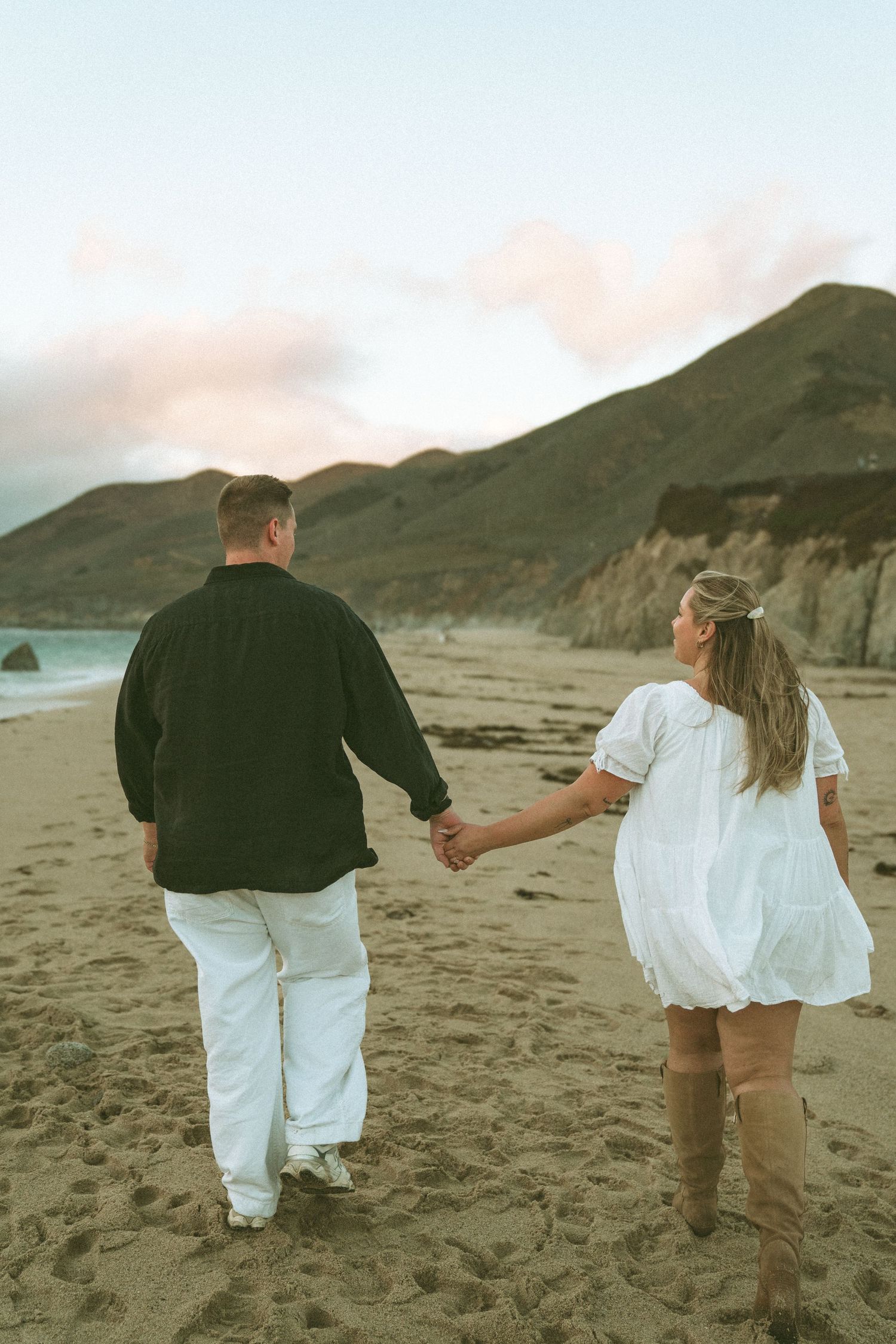 Couple holding hands while walking along a sandy beach with mountains and cliffs in the distance.