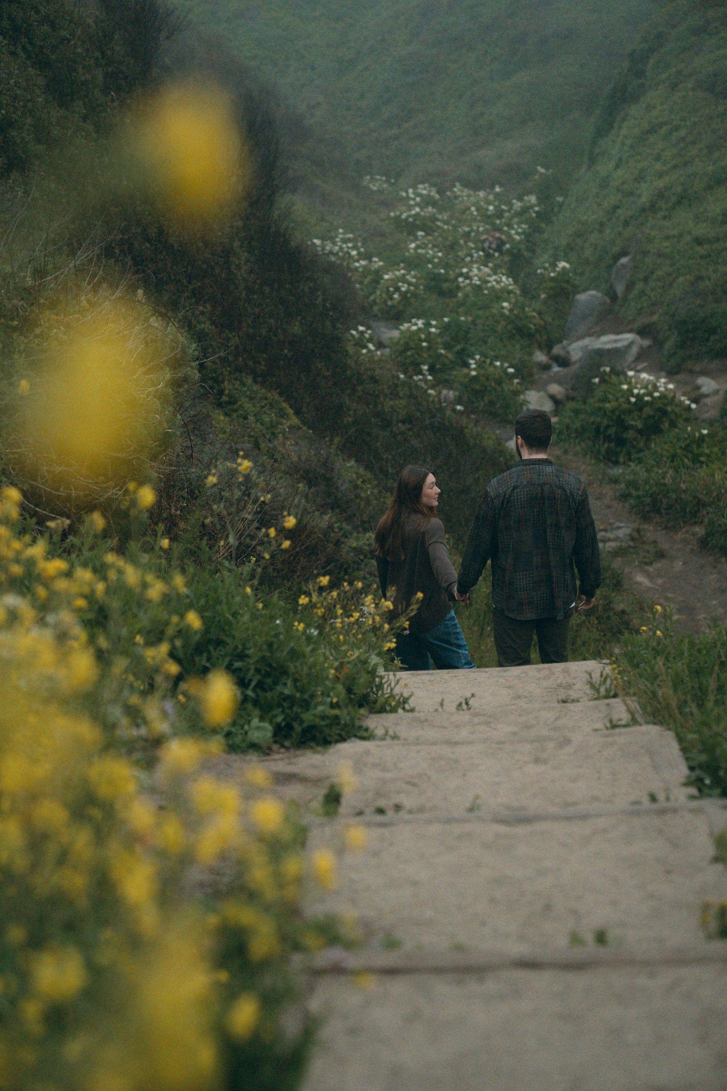 A couple walks down a mountain path surrounded by yellow wildflowers and lush green hillsides.
