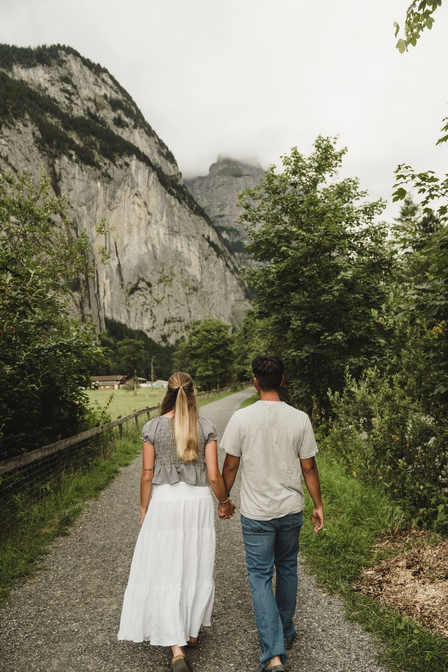 Couple walking hand in hand on mountain trail with towering cliff face and lush greenery in Switzerland.