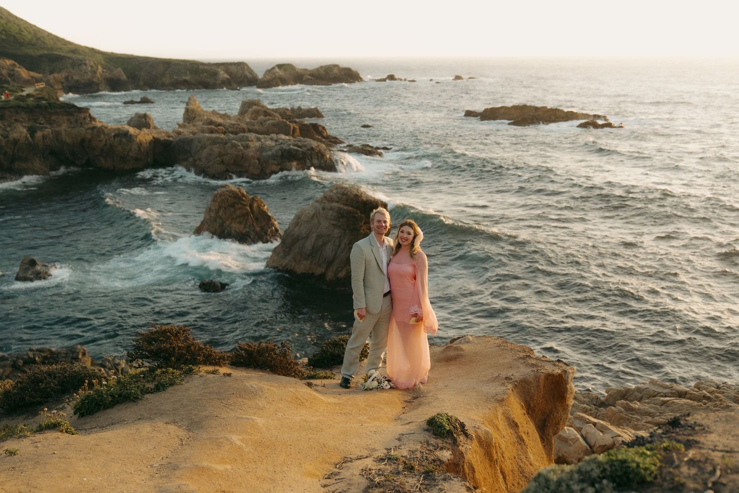Couple standing on golden coastal cliffs overlooking rocky Pacific Ocean shoreline at sunset in Big Sur, California.