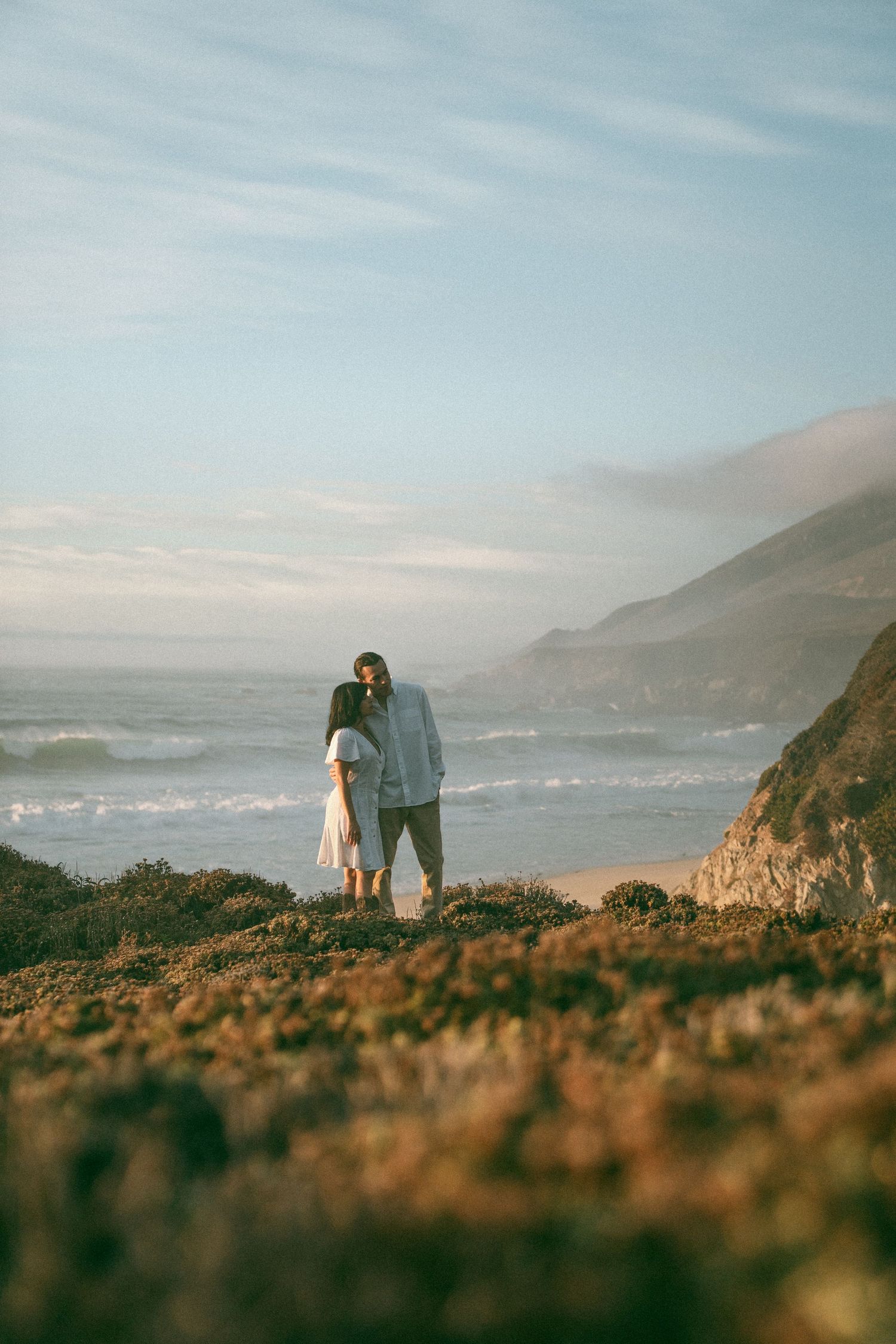 Two figures walk along a scenic coastal cliff path with dramatic ocean views and mountains in the distance.