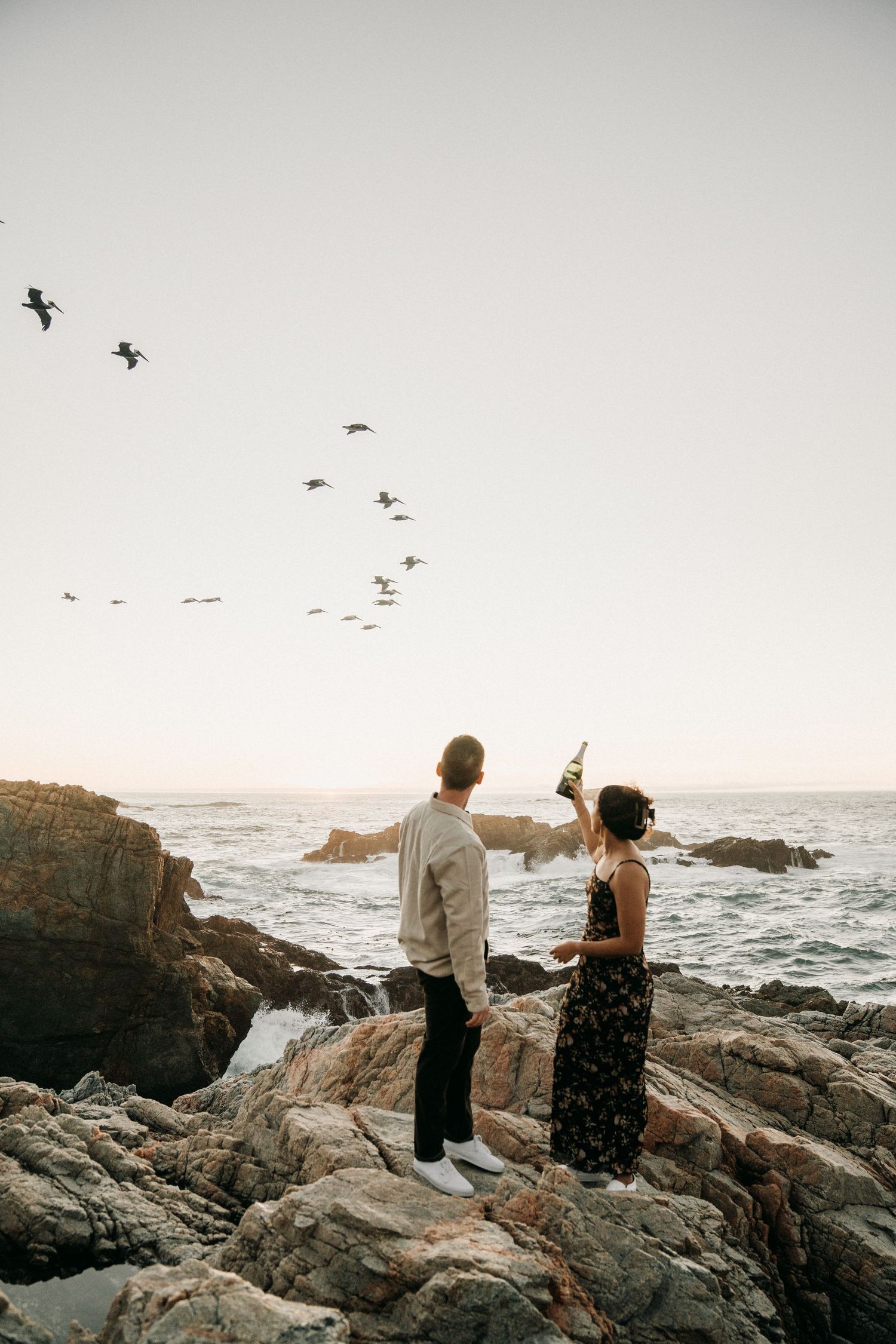 Couple enjoying sunset views on rugged oceanside cliffs with seabirds flying overhead.