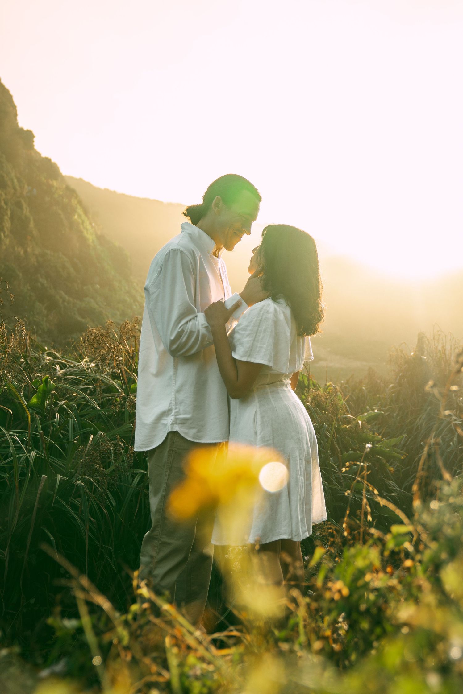 A couple in white clothing embraces tenderly in a sunlit meadow during golden hour as wildflowers sway in the breeze.