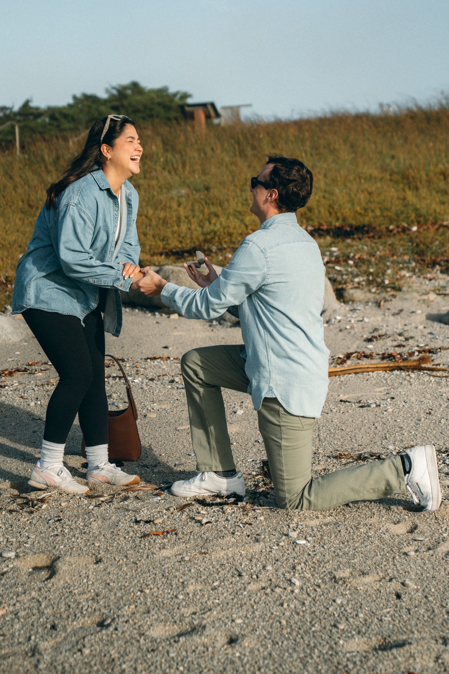 A proposal moment captured on a rocky beach with one person kneeling and another reacting with joy.