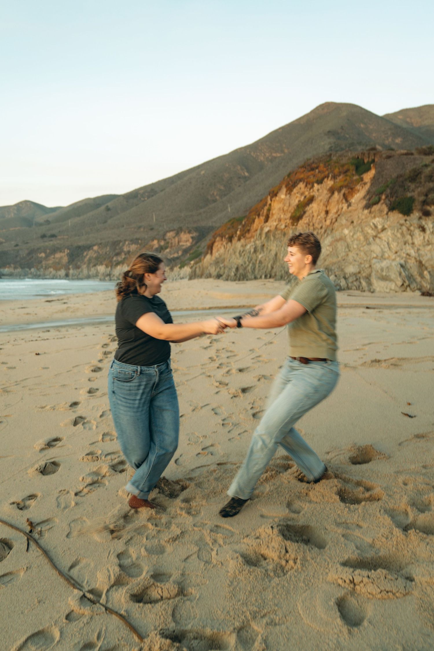 Playful couple pretending to push each other on sandy beach with cliffs in background.
