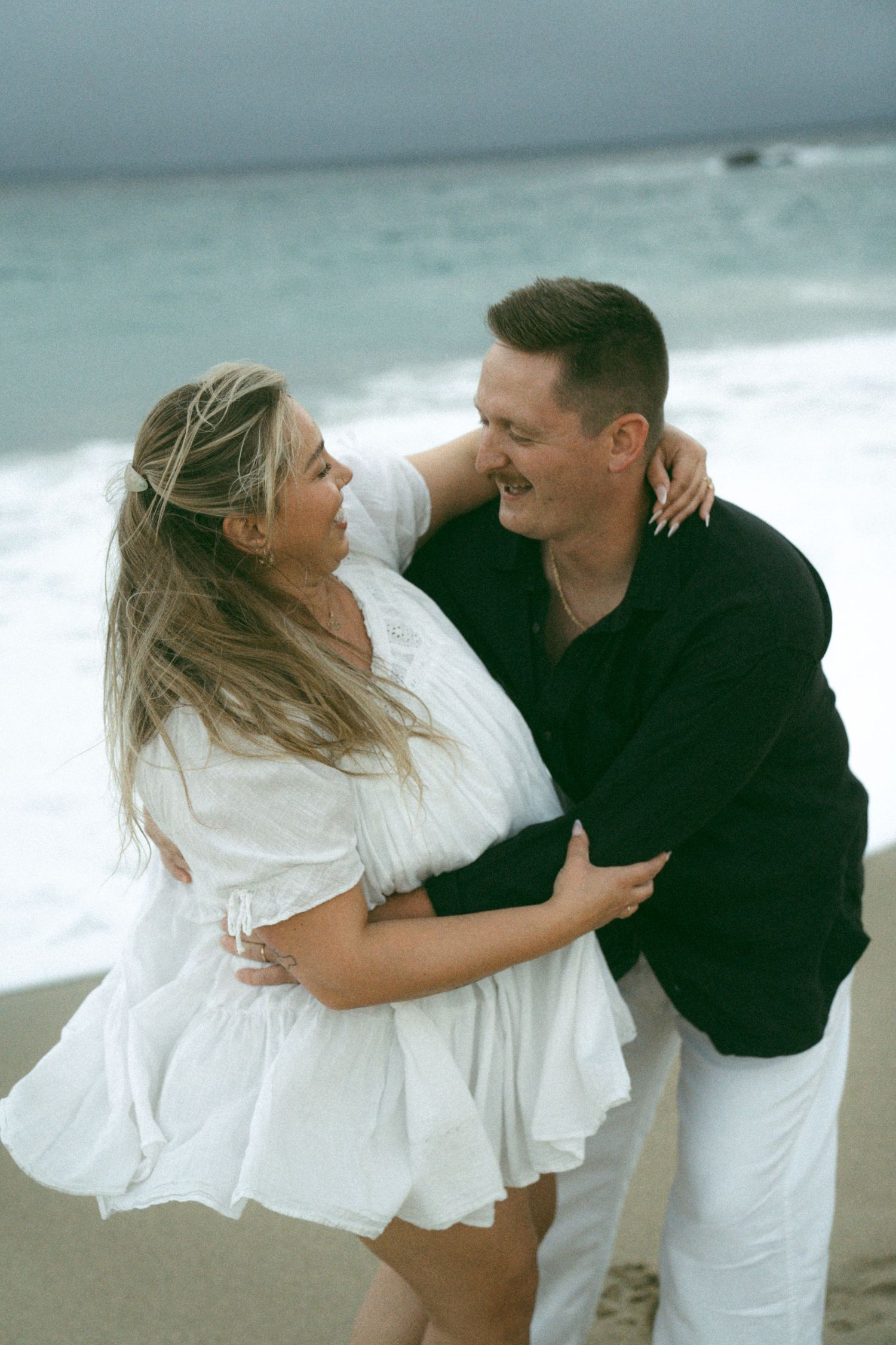 A couple in coordinating black and white outfits embrace playfully on a misty beach shoreline.