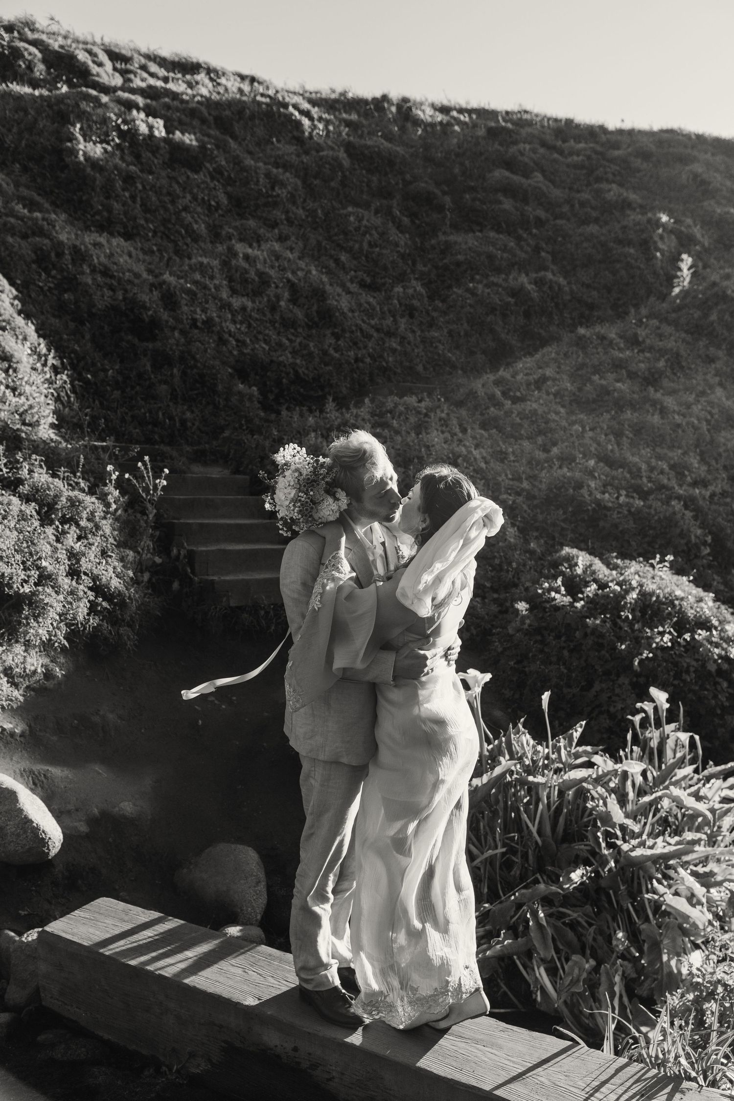 Black and white photo of a couple kissing on a wooden deck surrounded by lush green hills on their wedding day.